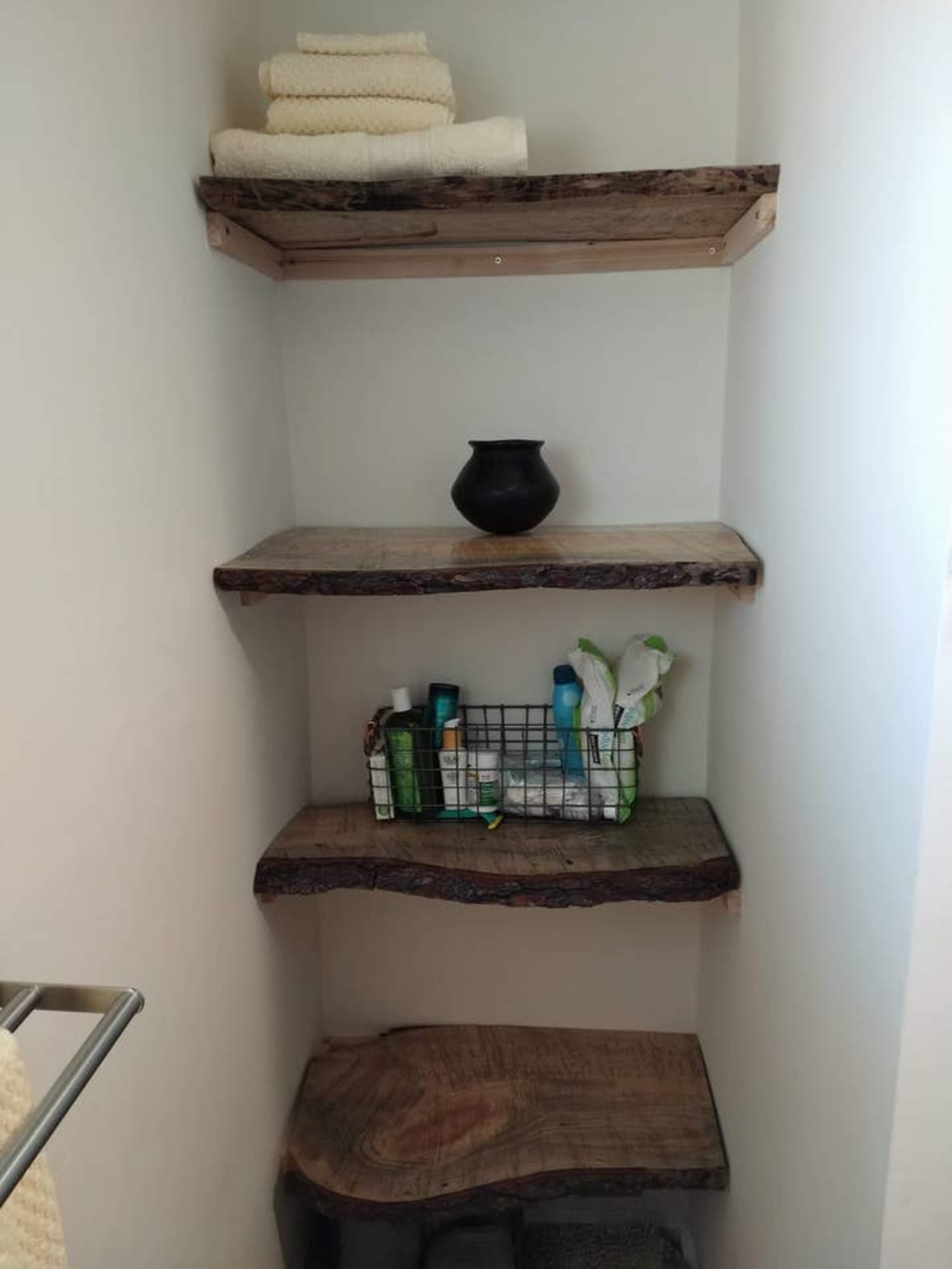 A corner shelving unit with three wooden shelves displaying neatly stacked towels, a black vase, and various personal care items in a wire basket.