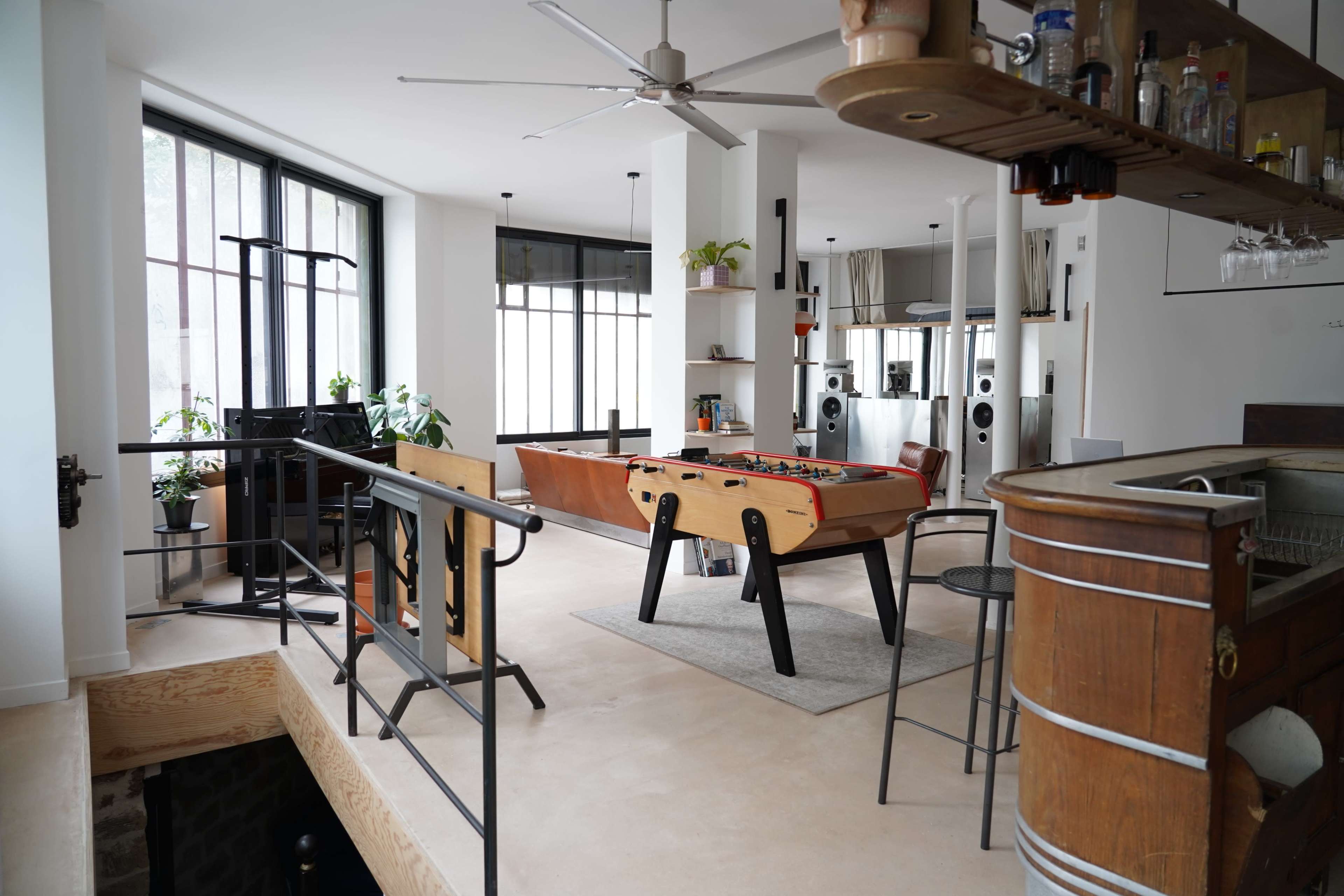 The image shows a modern open-concept living space featuring a game table in the center, surrounded by large windows and minimalist décor.