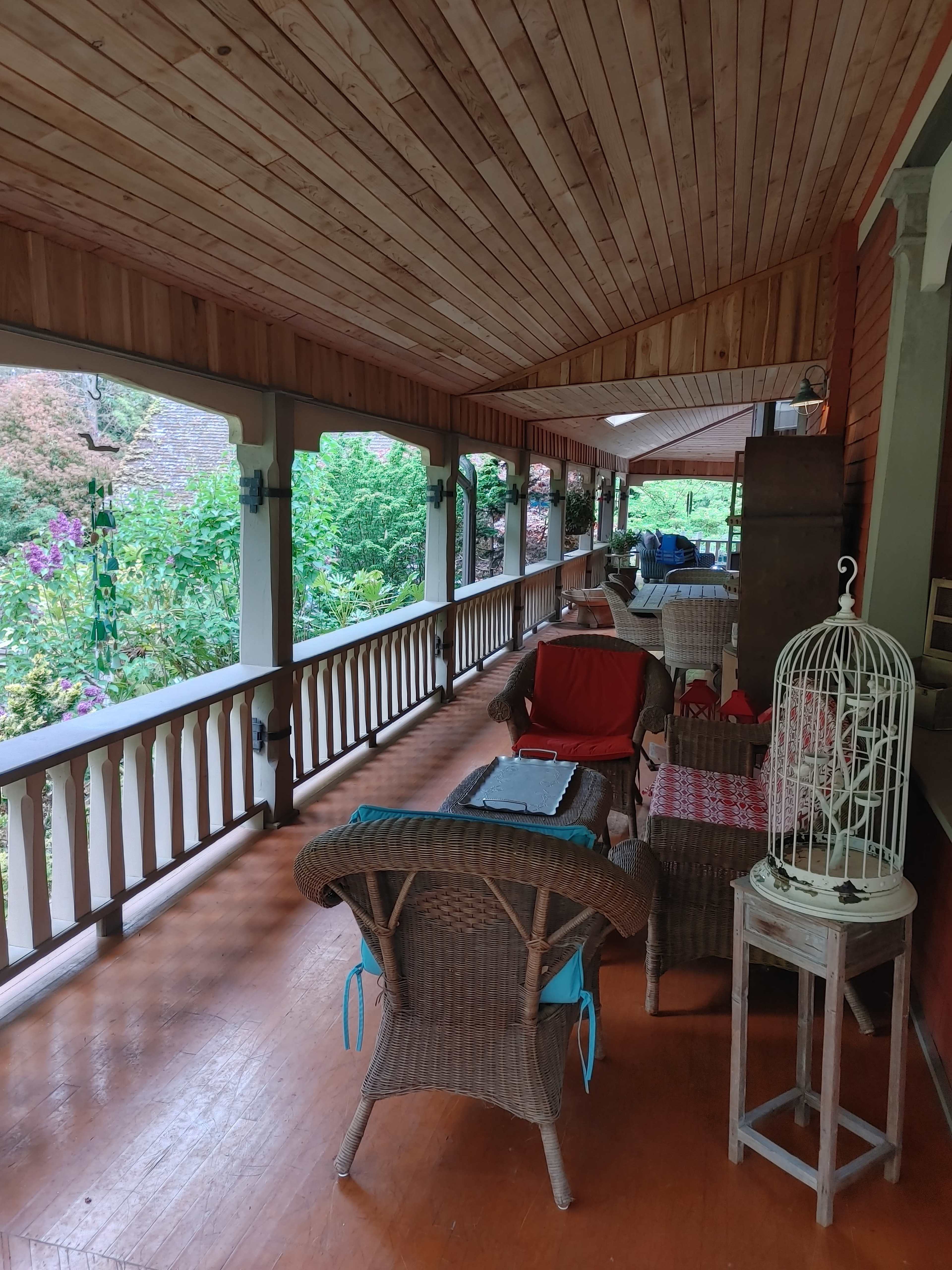 A spacious porch with wicker furniture, a white birdcage, and greenery visible through the railings.