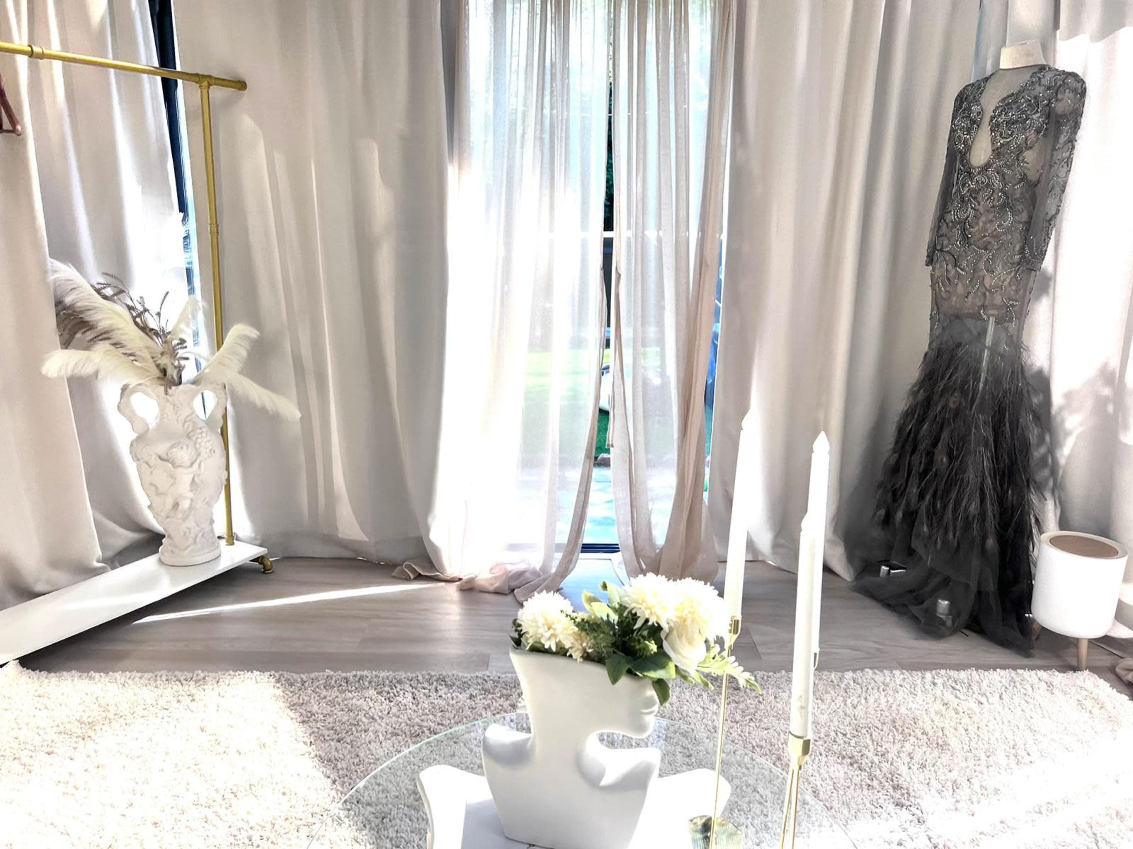 The scene features a elegantly decorated room with a floor-length evening gown on a mannequin, a vase with flowers, and a glass table with candles, all set against sheer curtains and a view of the outdoors.
