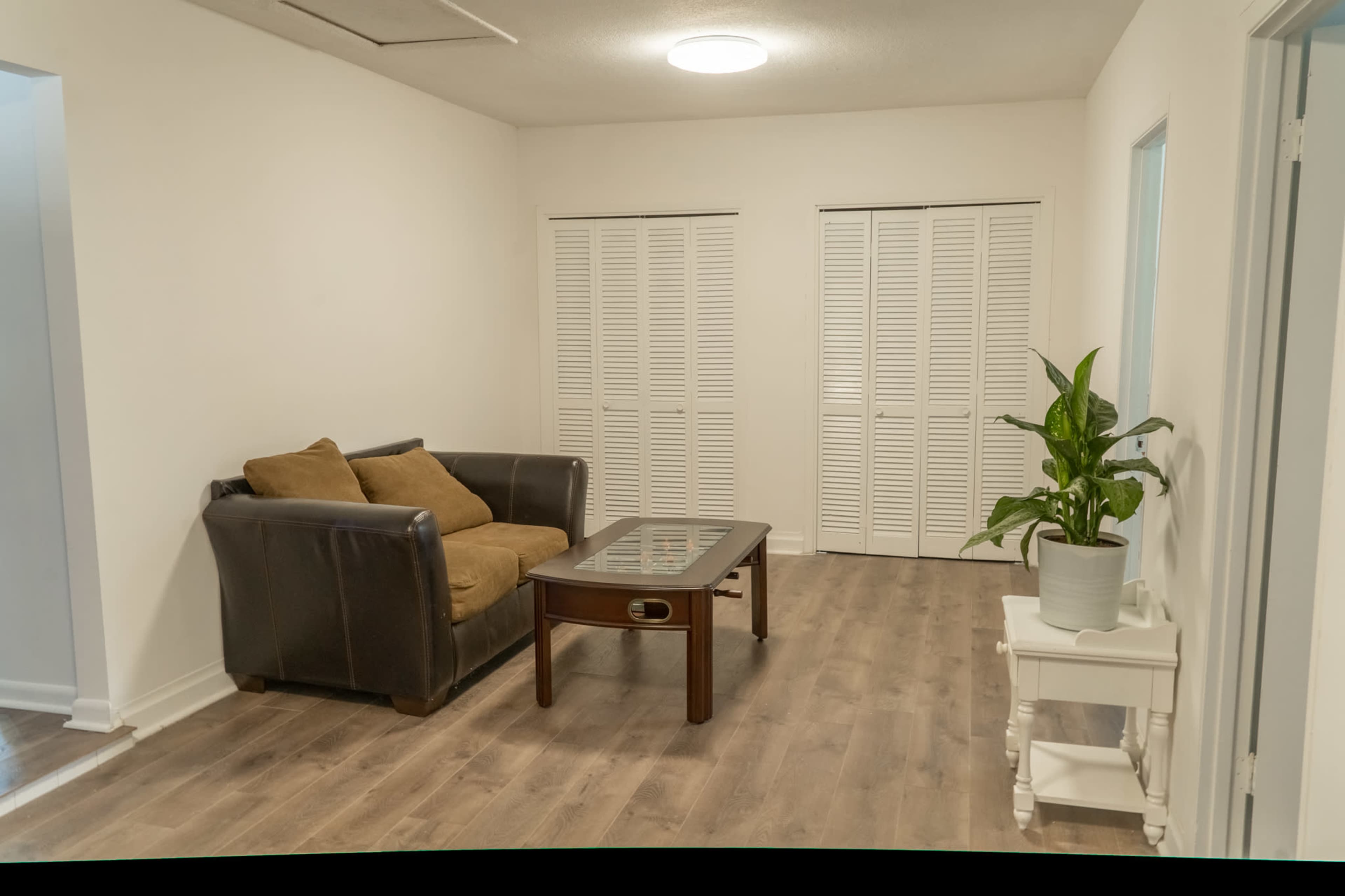 A simple living room featuring a brown couch, a glass coffee table, a potted plant, and white folding closet doors.