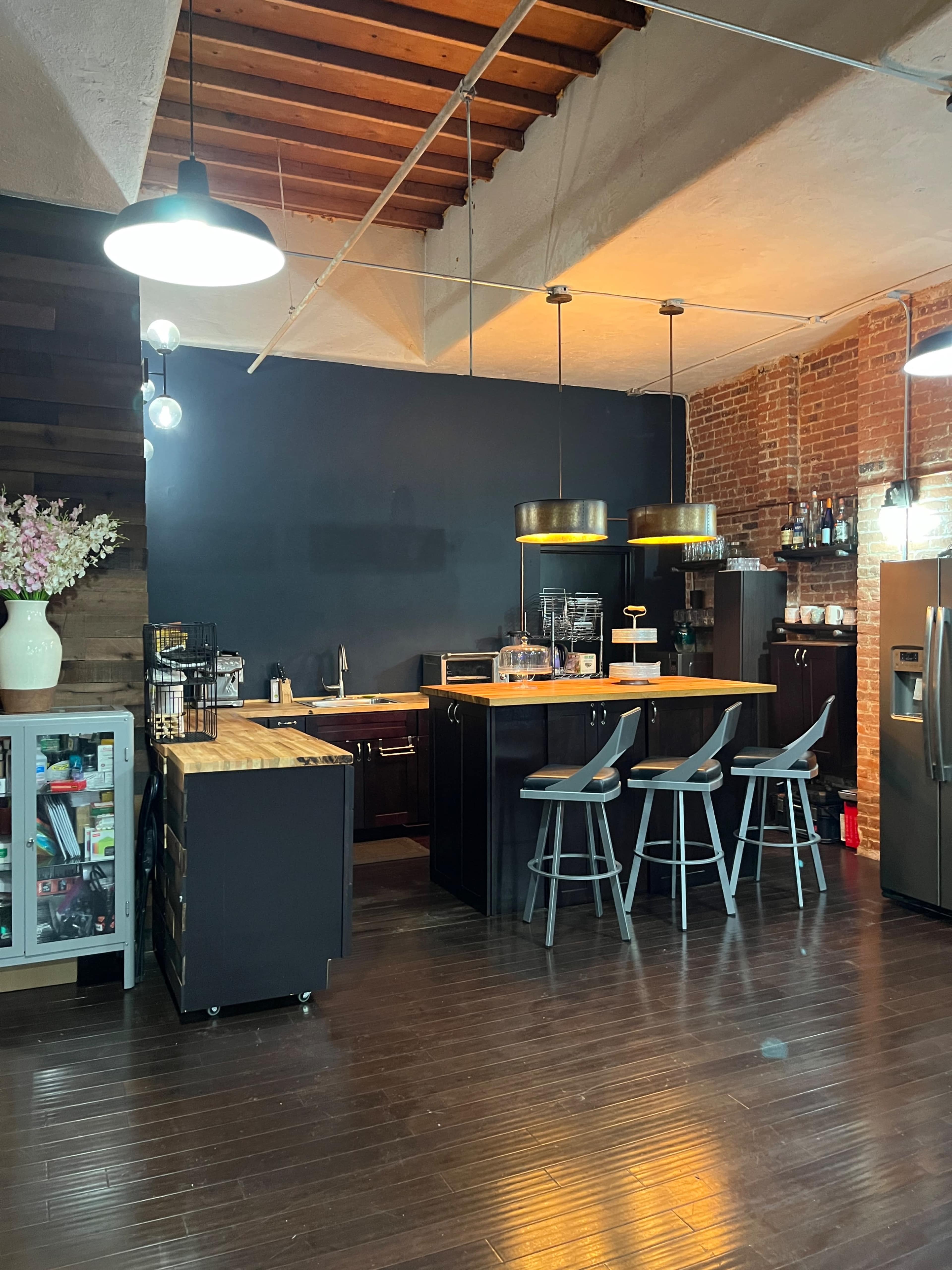 A modern kitchen with a wooden countertop, bar stools, and a mix of brick and dark painted walls.