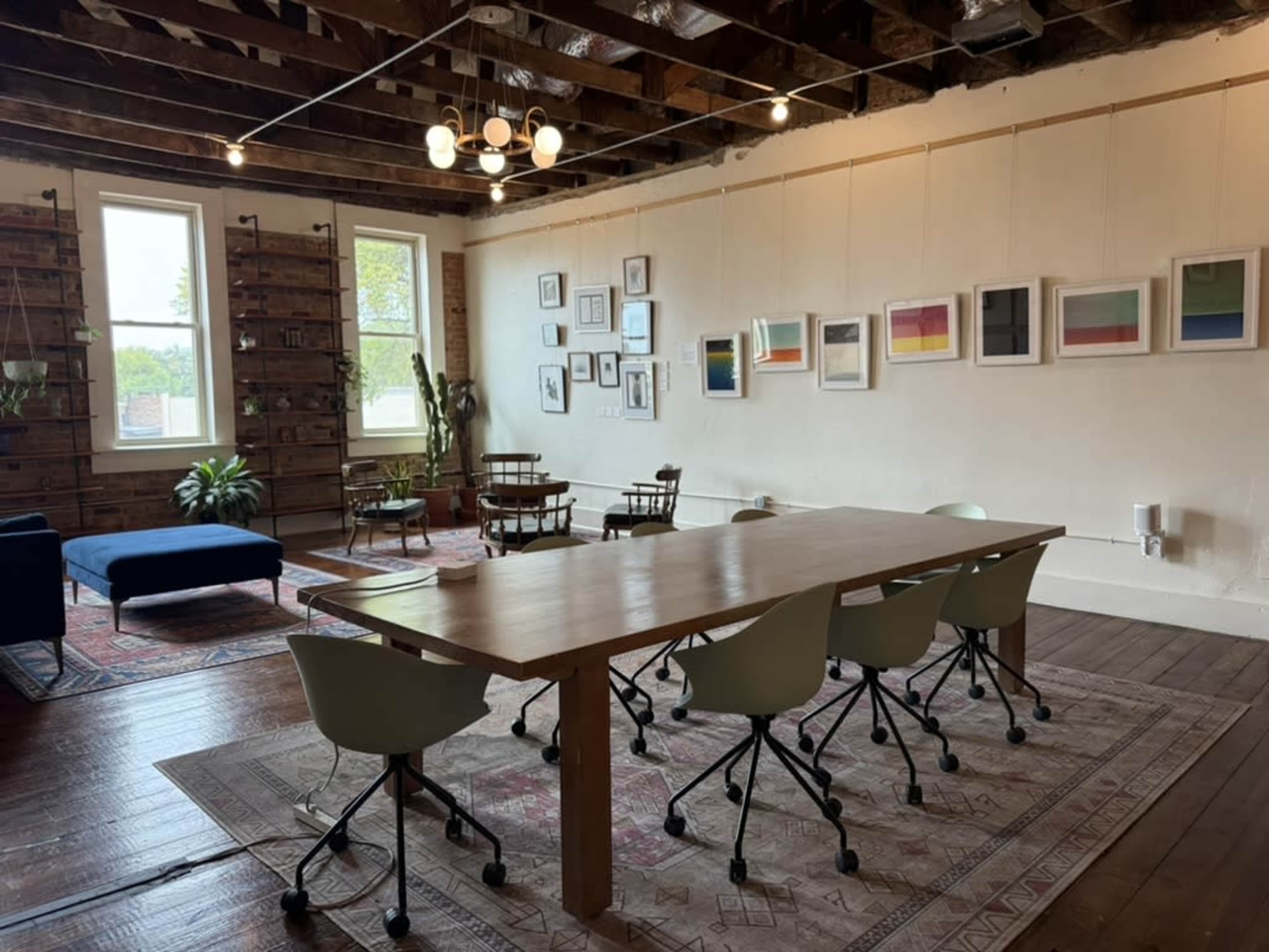 The image shows a spacious, well-lit room with wooden beams, a large table surrounded by chairs, and artworks displayed on the walls.