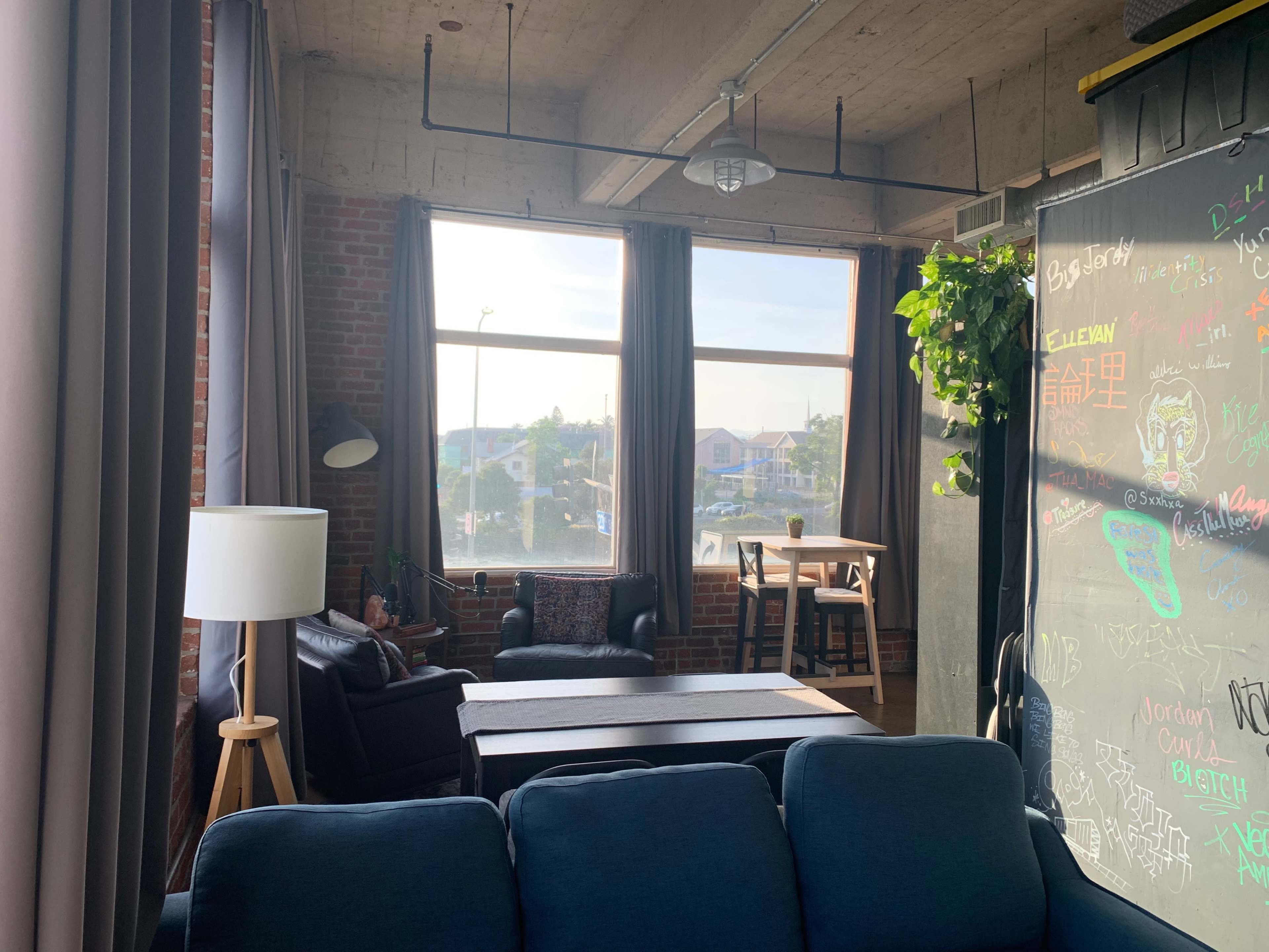A spacious living area with large windows, a sofa, a lamp, and a dining table, surrounded by brick walls and green plants.