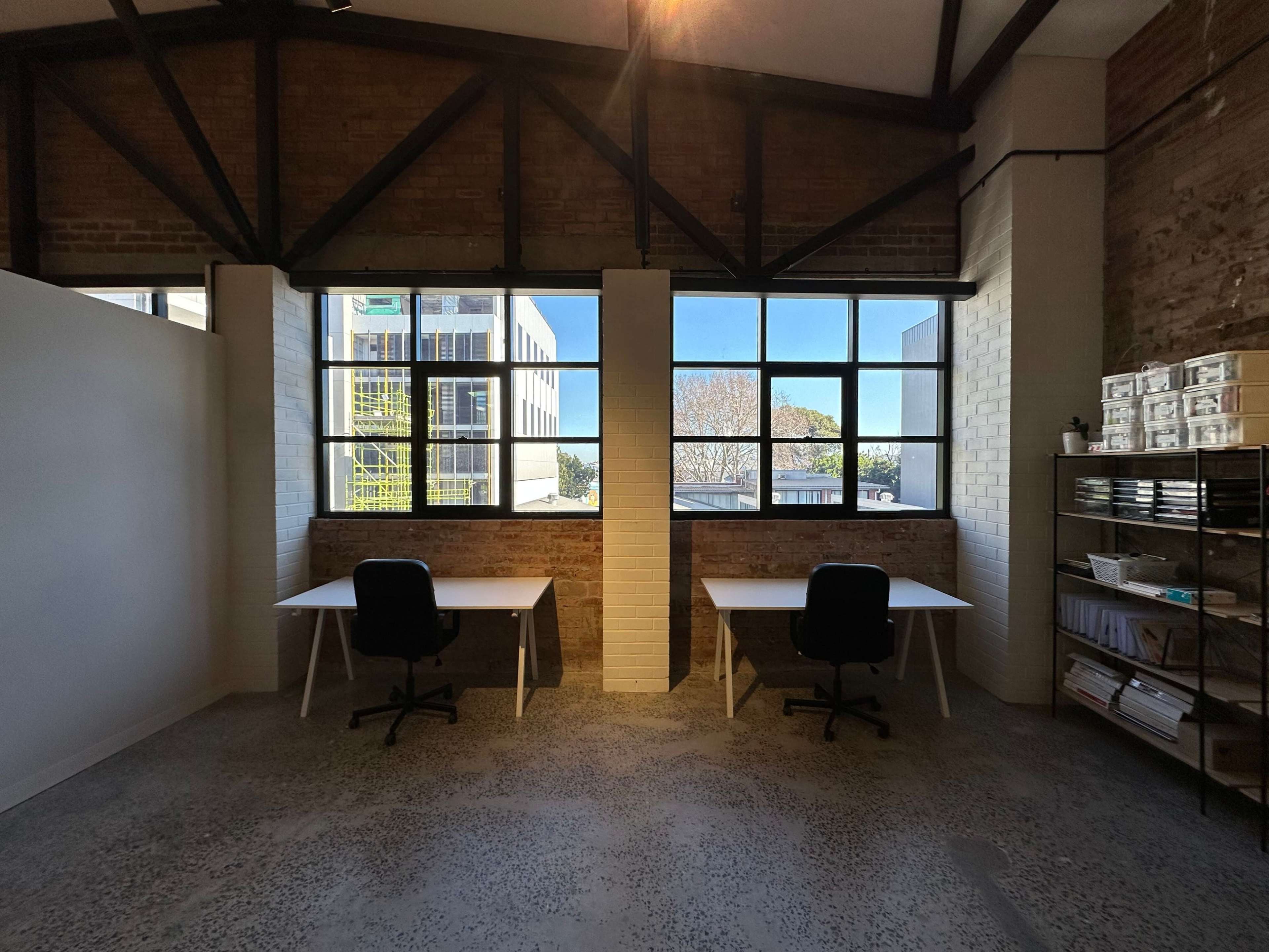 A spacious office interior with two desks and chairs positioned under large windows, framed by exposed brick walls and industrial-style beams.