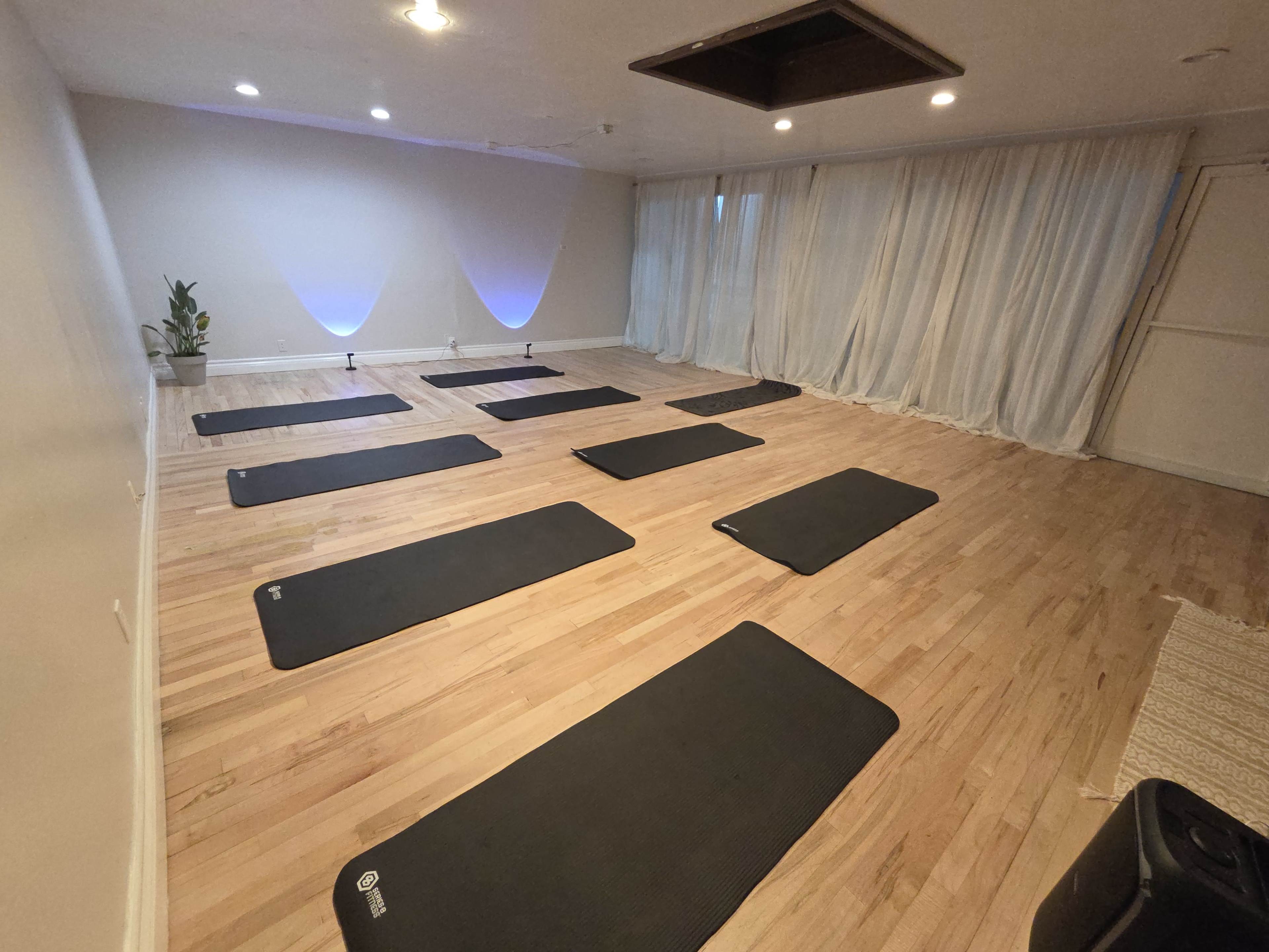 The image shows a yoga studio with several exercise mats arranged on a wooden floor, illuminated by soft lighting and surrounded by sheer curtains.