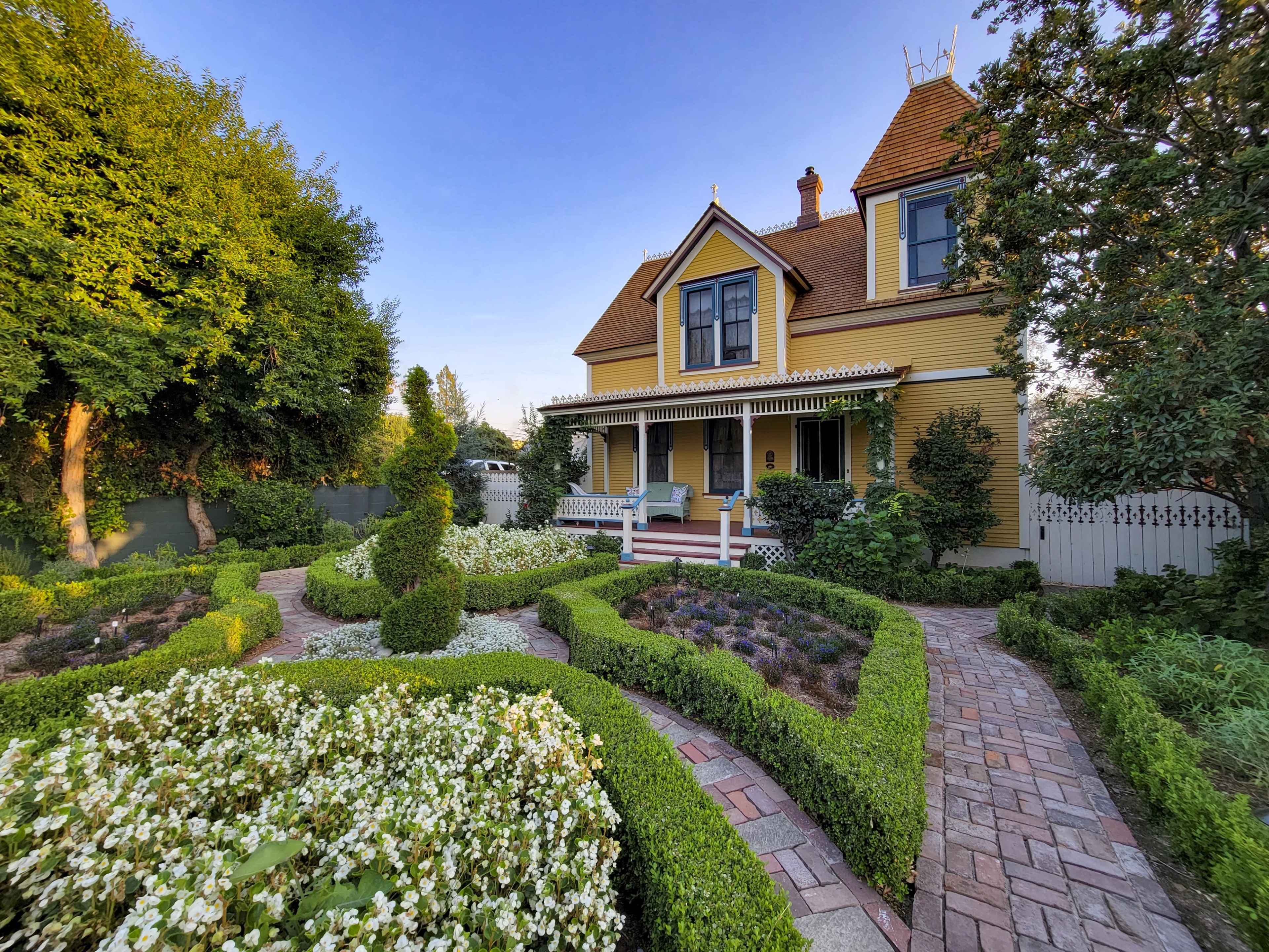 A yellow Victorian-style house with a steep roof is surrounded by well-manicured gardens and pathways.