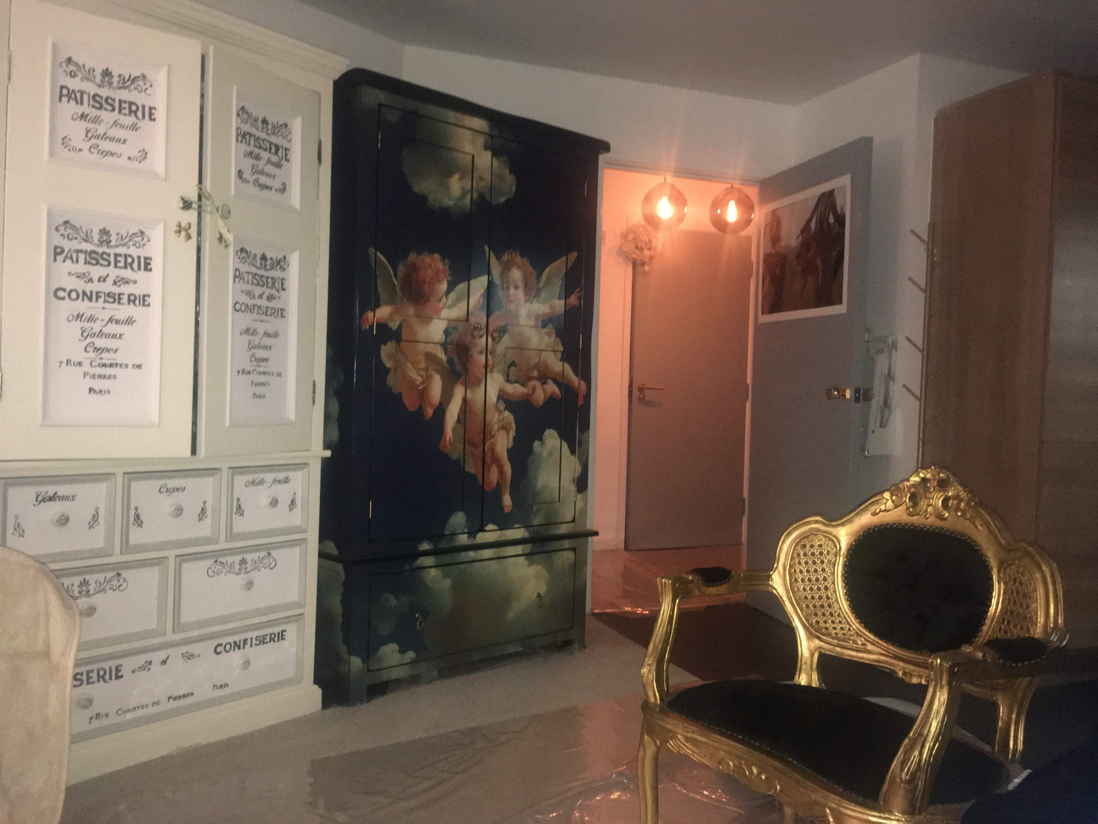 The image shows a room with a large, painted cabinet featuring cherubs, an ornate gold chair, and various decorative items on the walls.