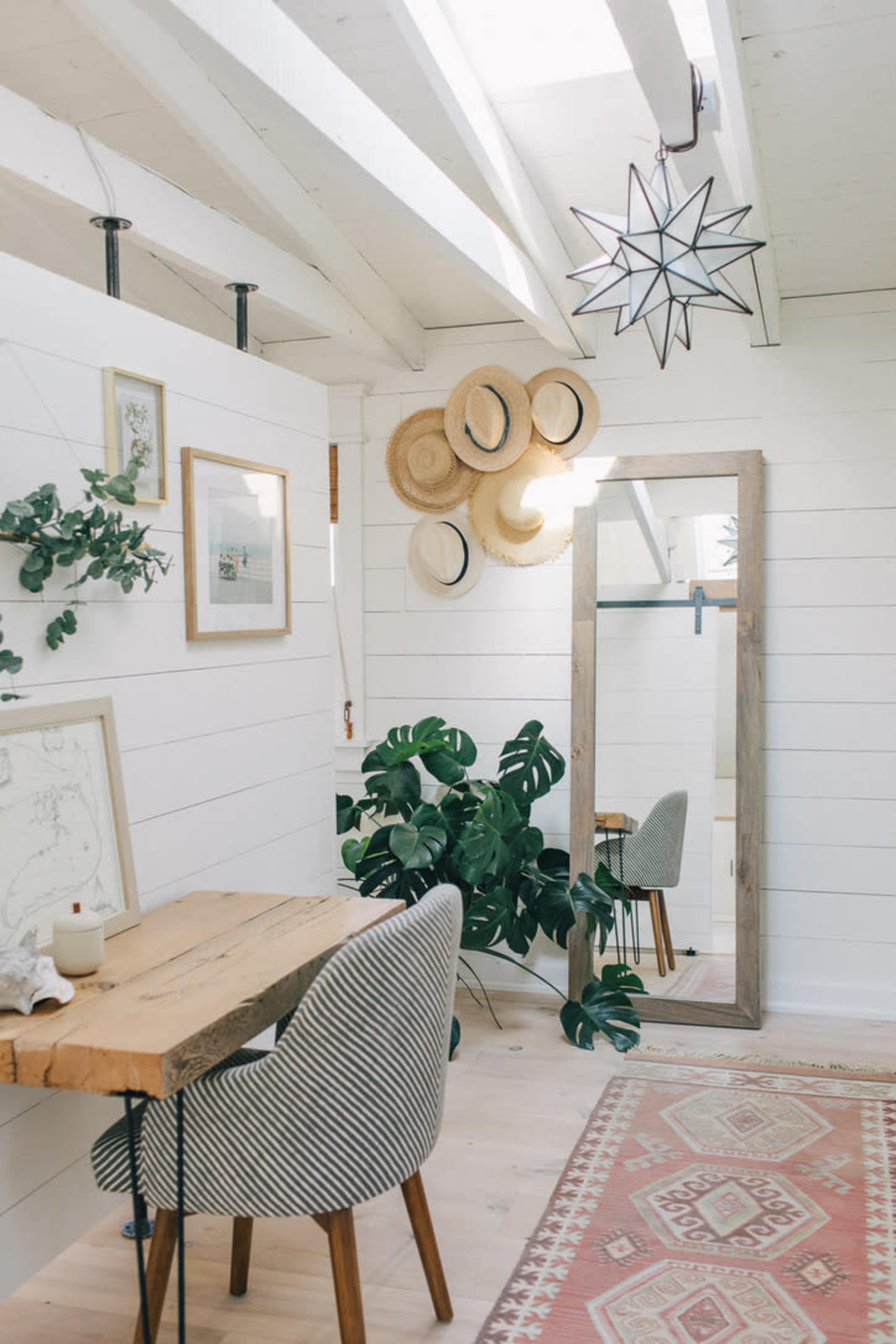 The image shows a bright, minimalist room with a wooden table and chair, a mirror, decorative wall art, and greenery, accented by a star-shaped light fixture and round straw hats.