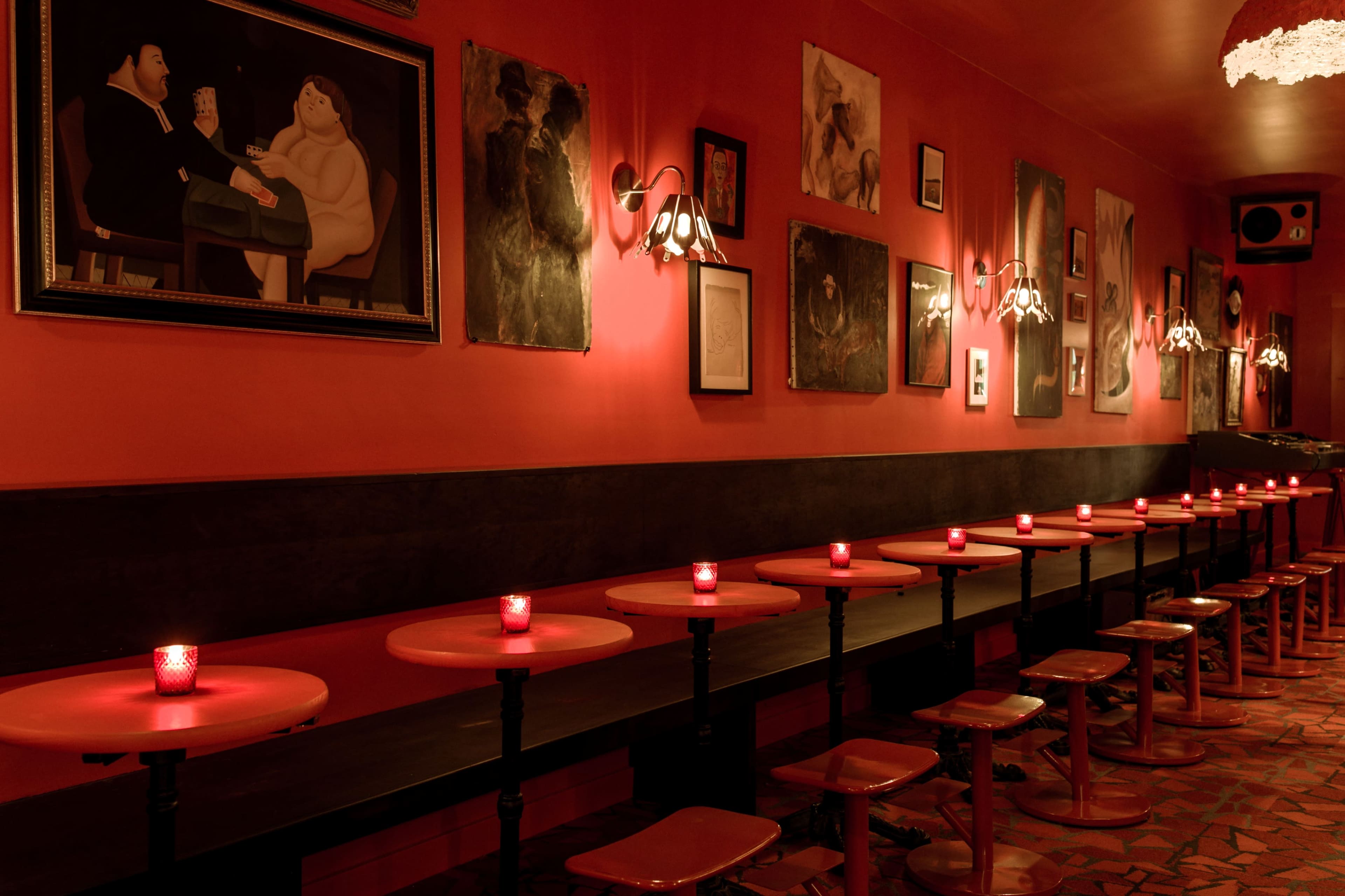 A dimly lit bar features a row of small round tables with red candles and stools, against a backdrop of red walls adorned with various paintings.