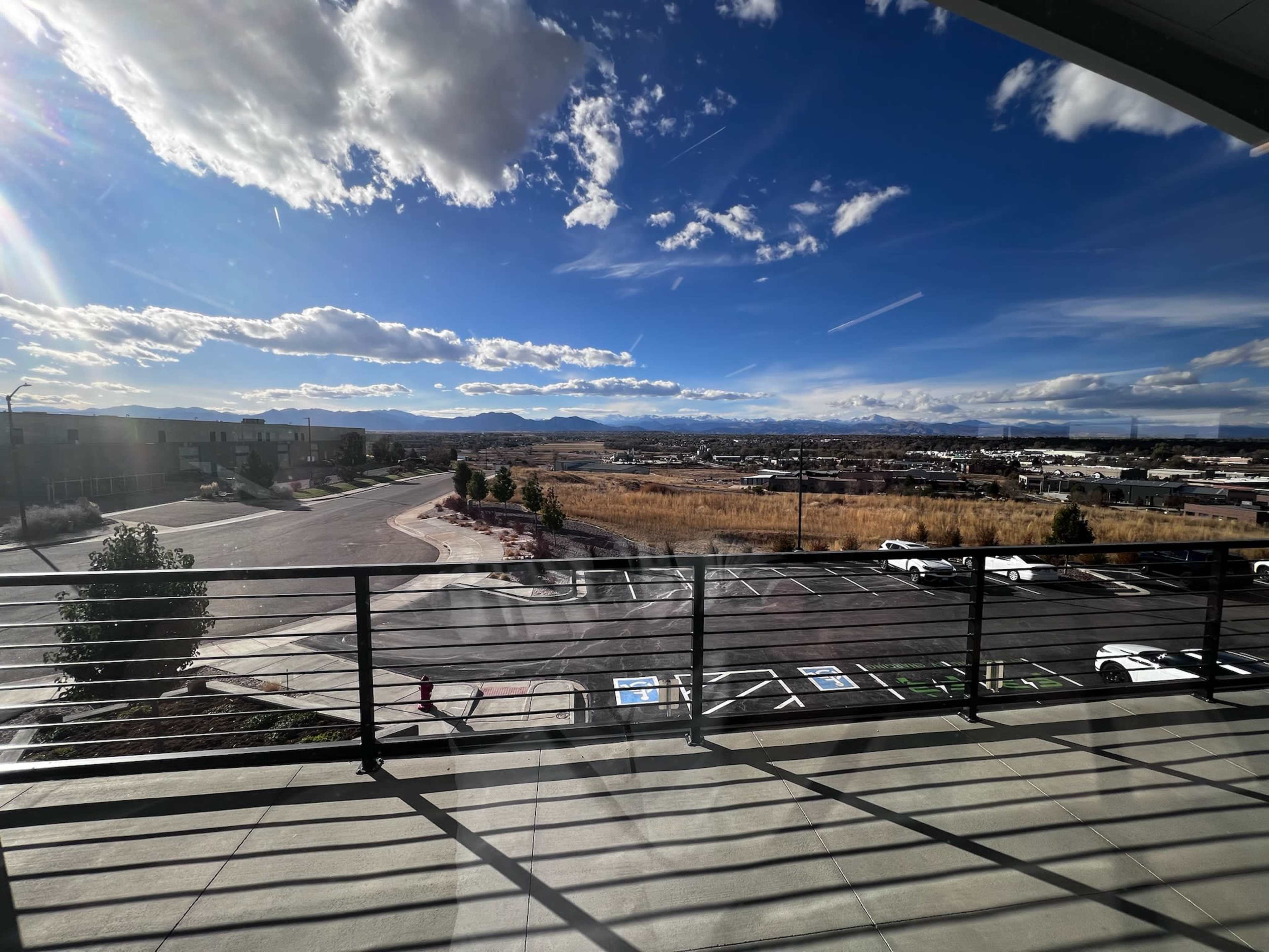A view from a balcony overlooking a road, parking area, and distant mountains under a partly cloudy sky.