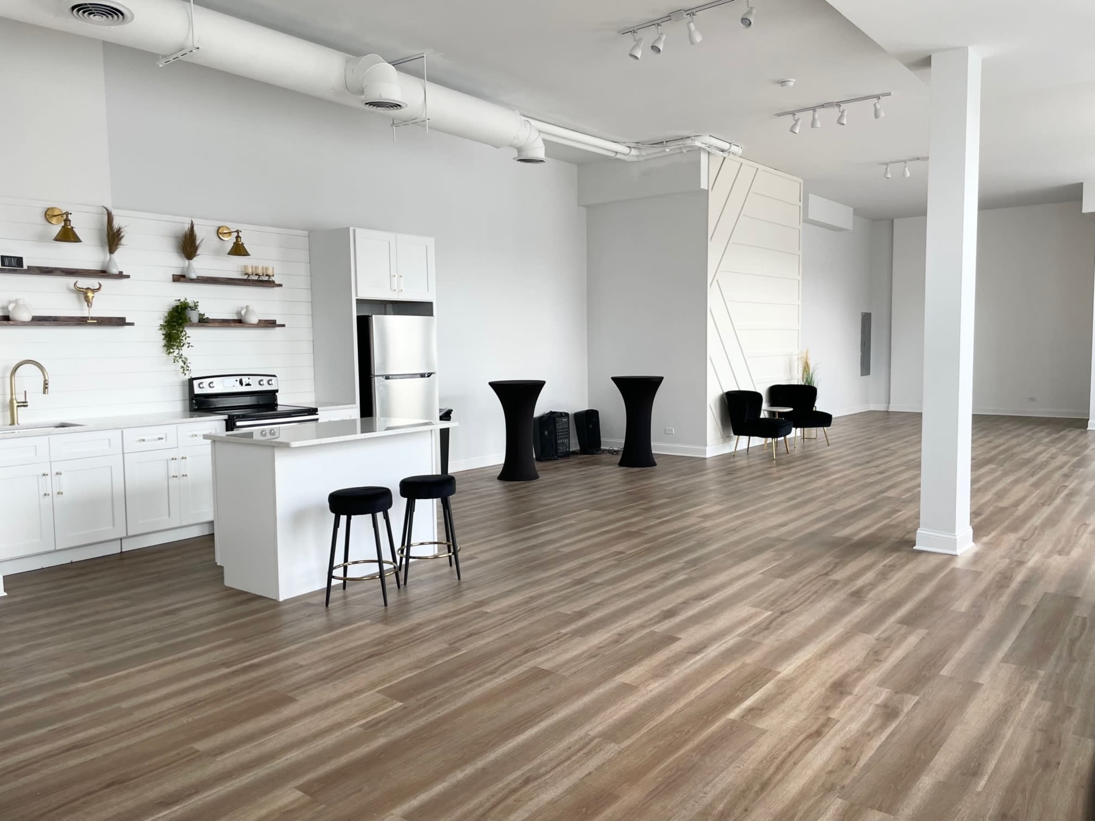 A spacious, modern interior with a kitchenette on one side and black seating arrangements against a white wall.