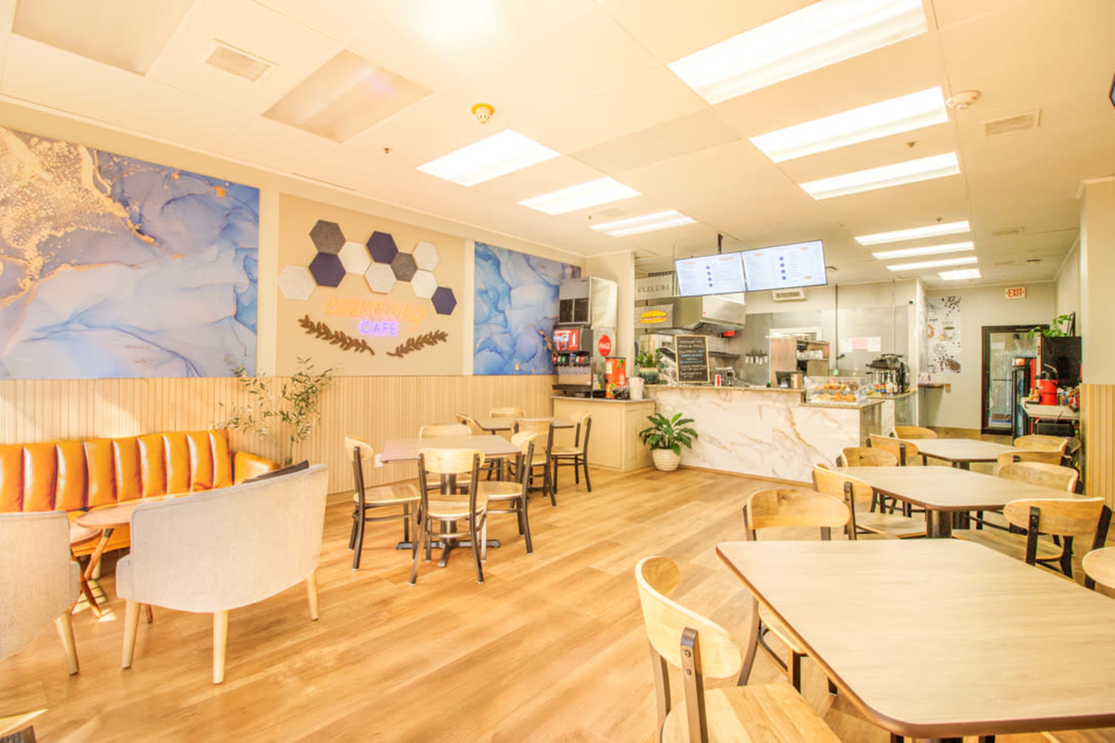 The image shows a cozy café interior with a mix of seating options, including chairs and tables, and a service counter displaying a menu.