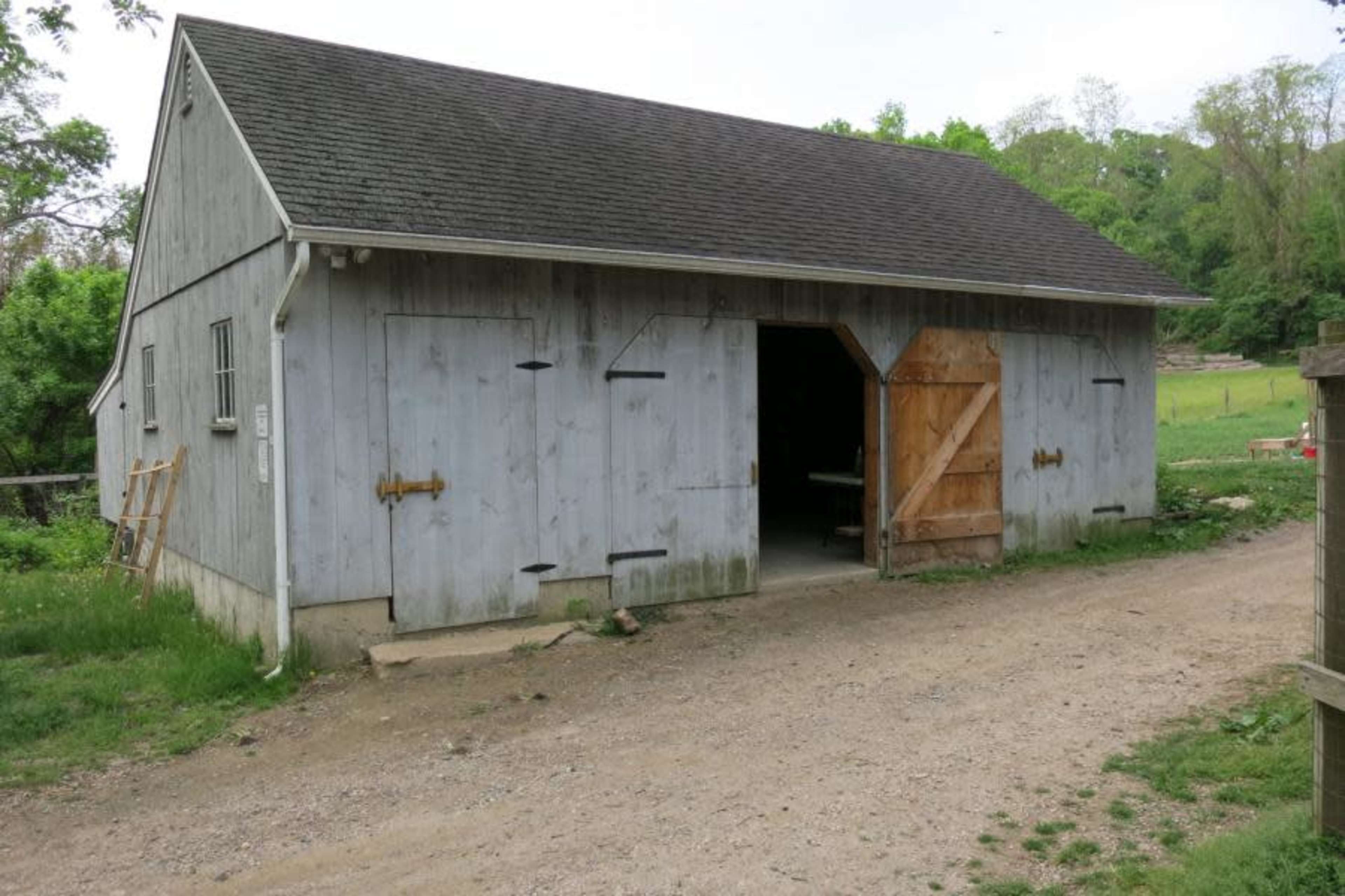 A large, gray wooden barn with open doors sits along a gravel path surrounded by grass and trees.
