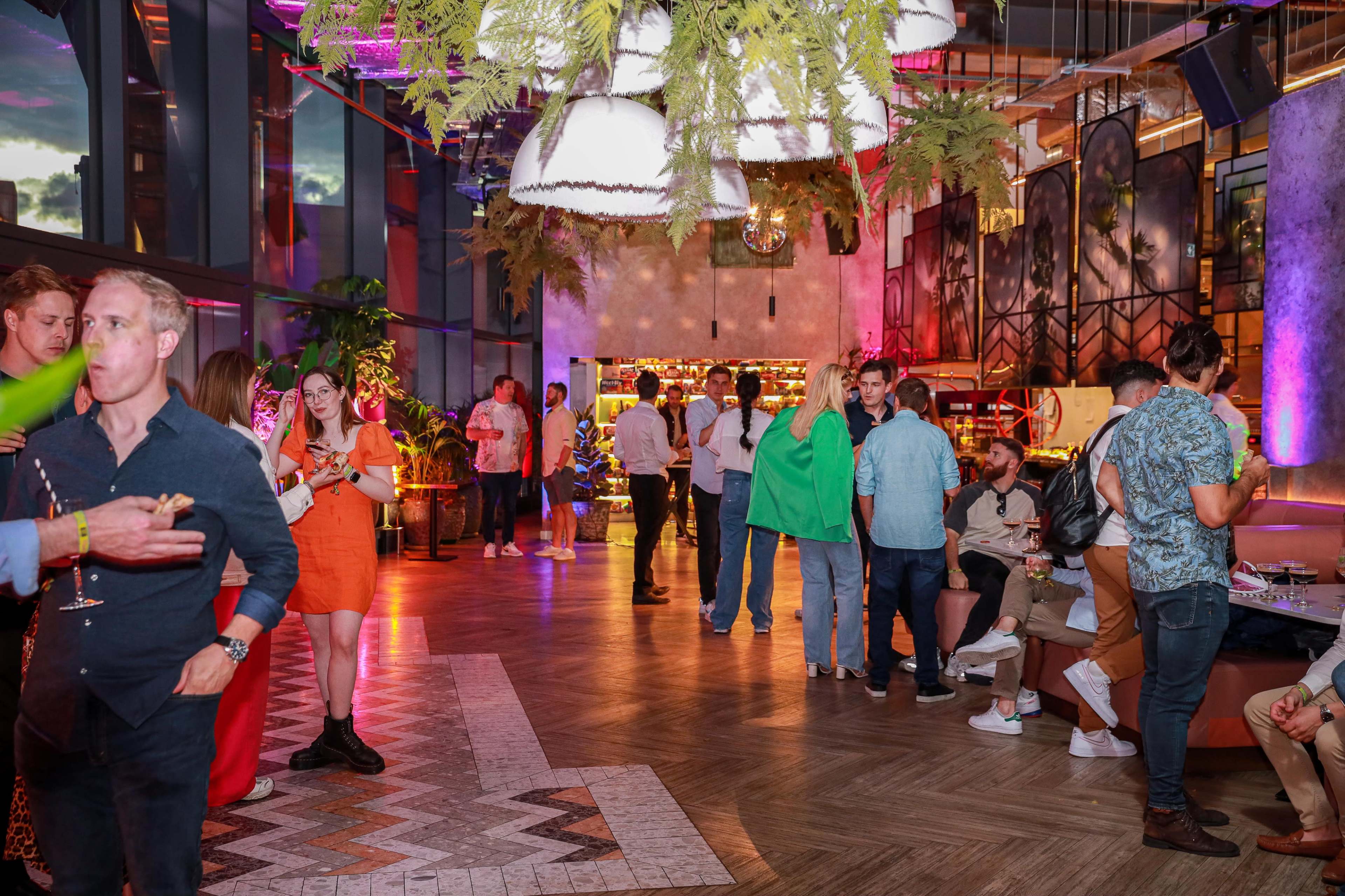 A lively bar scene features groups of people socializing under vibrant lighting, with a stylish decor that includes hanging plants and modern furniture.