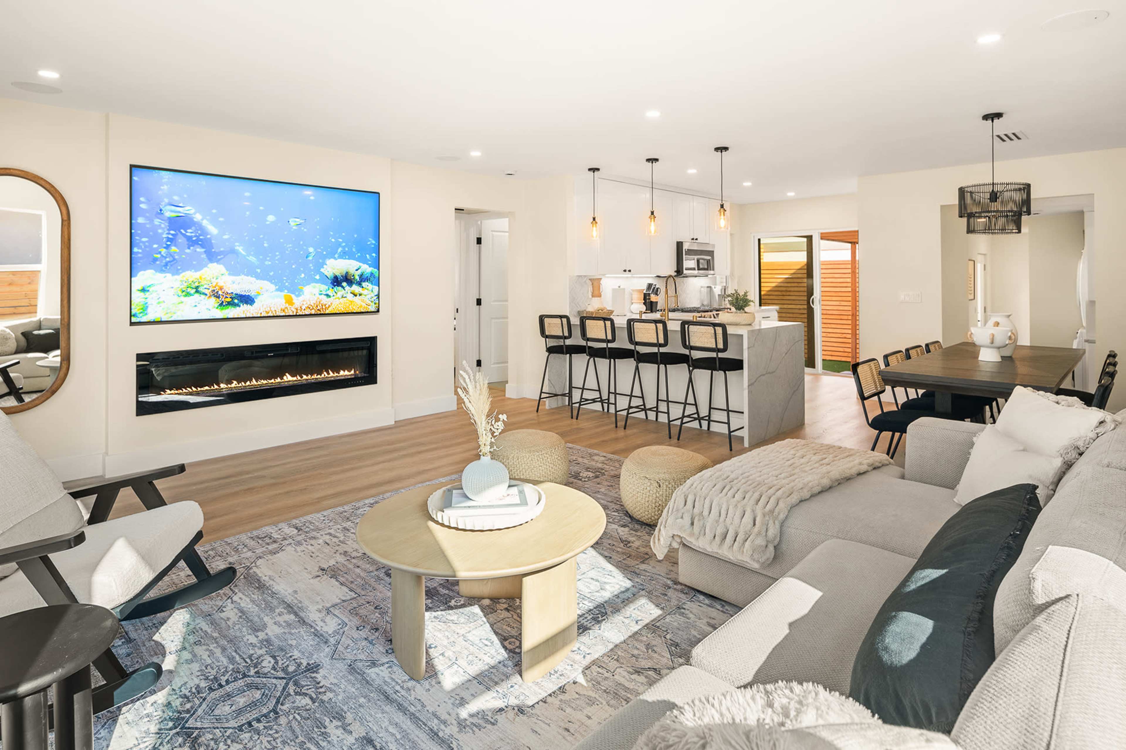 The image shows a modern living room featuring a large television mounted on the wall above a sleek gas fireplace, with a sectional sofa, a round coffee table, and an adjacent dining area with bar stools and a kitchen.