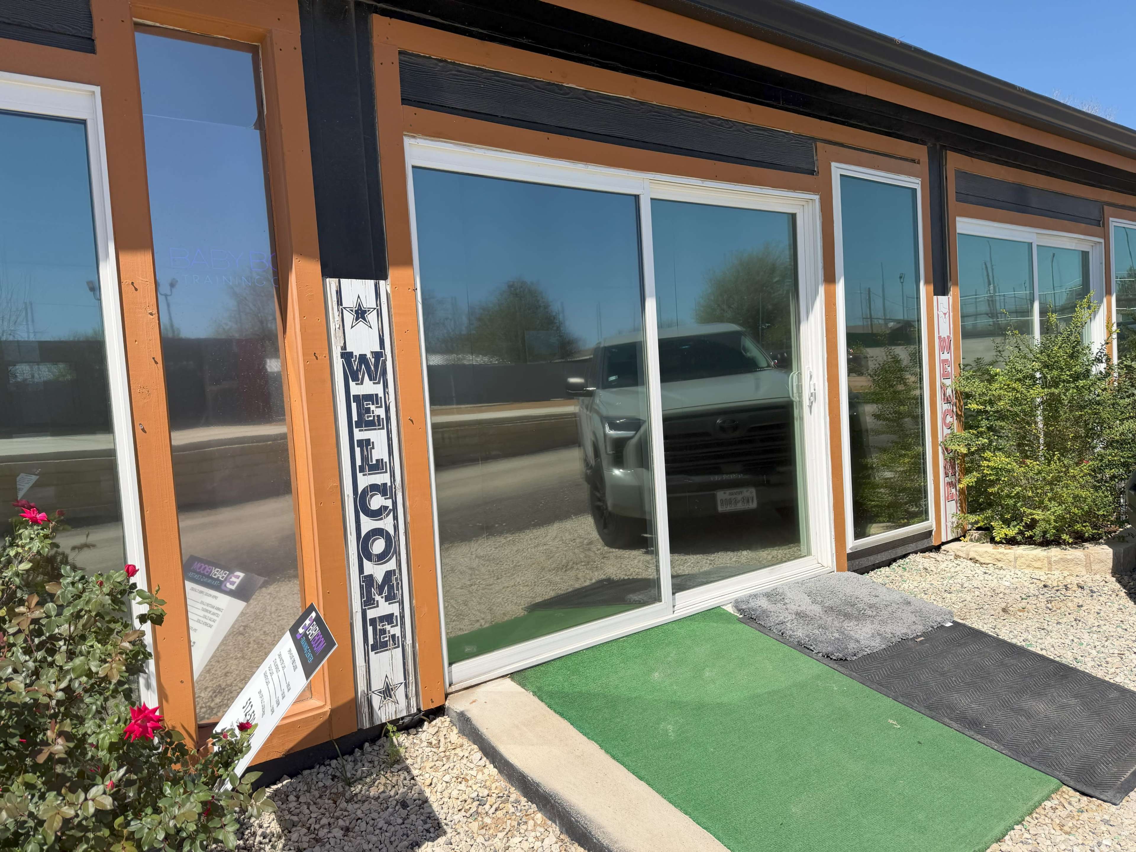 The image shows a building entrance with large glass doors, a "WELCOME" sign on the left, and a small pathway leading up to the entrance.