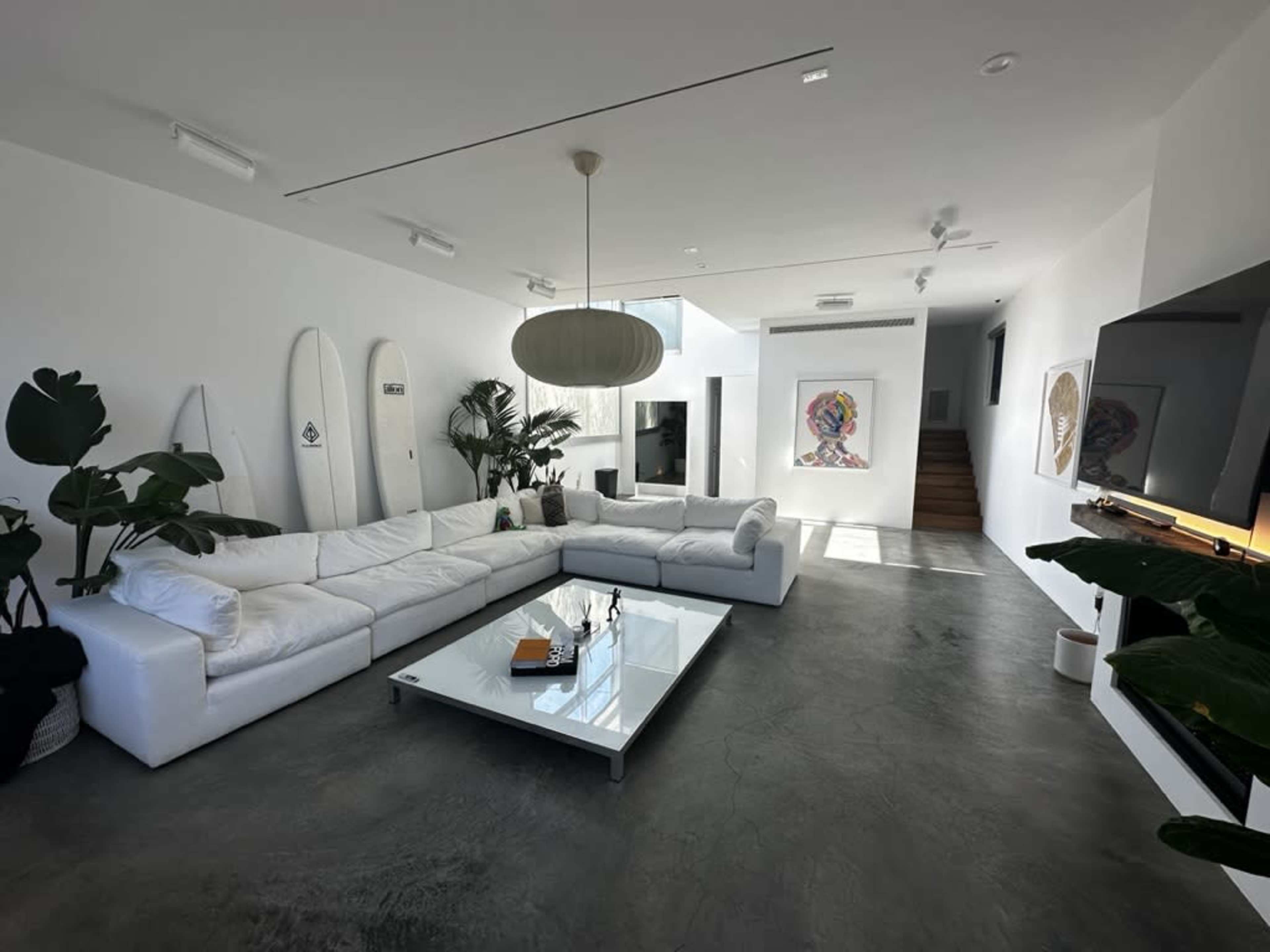 A modern living room with a large white sectional sofa, a glass coffee table, and indoor plants, set against a minimalist white wall and a staircase in the background.