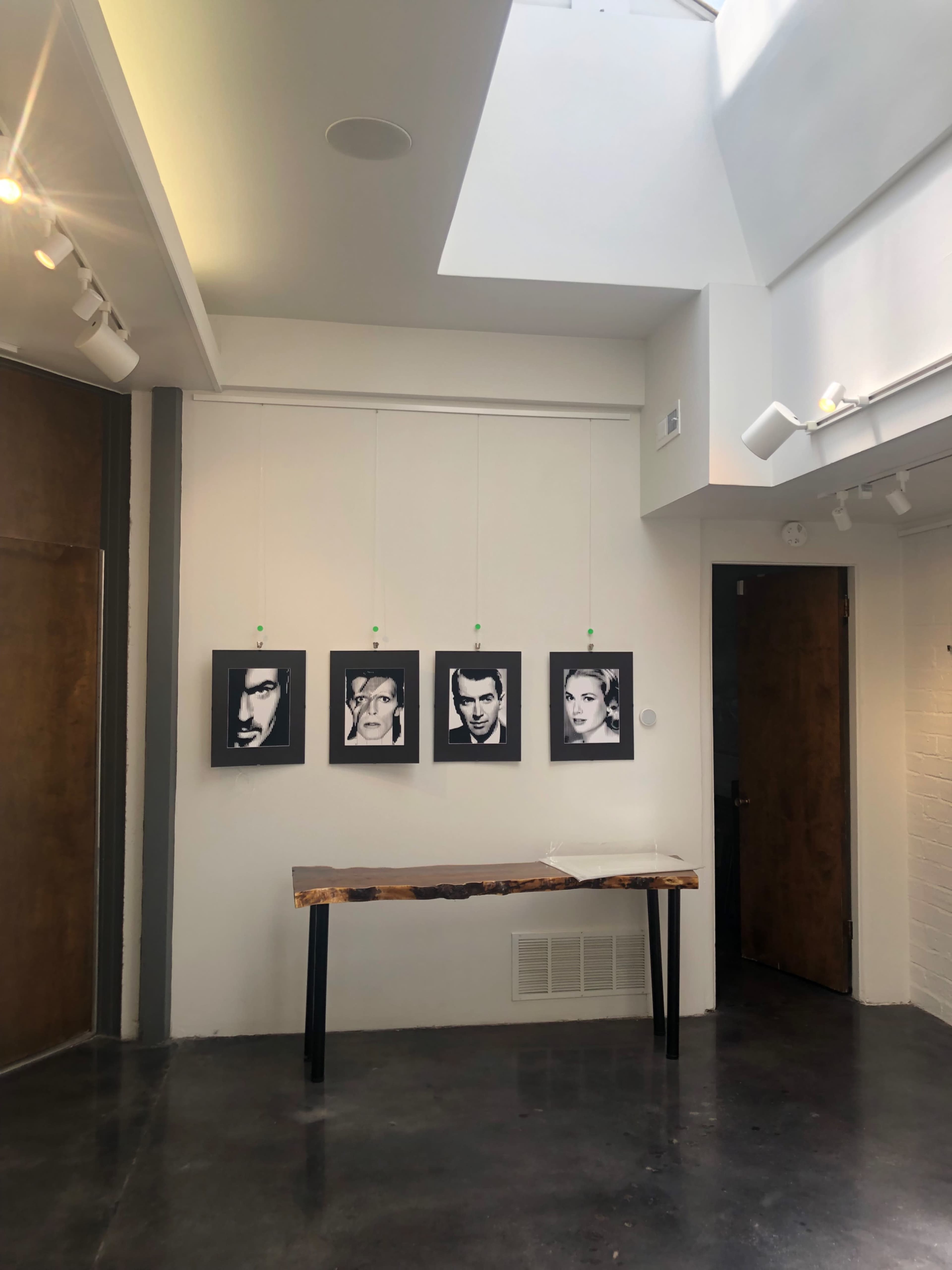 The image shows a gallery space featuring four black-and-white framed portraits hanging on the wall above a wooden table.
