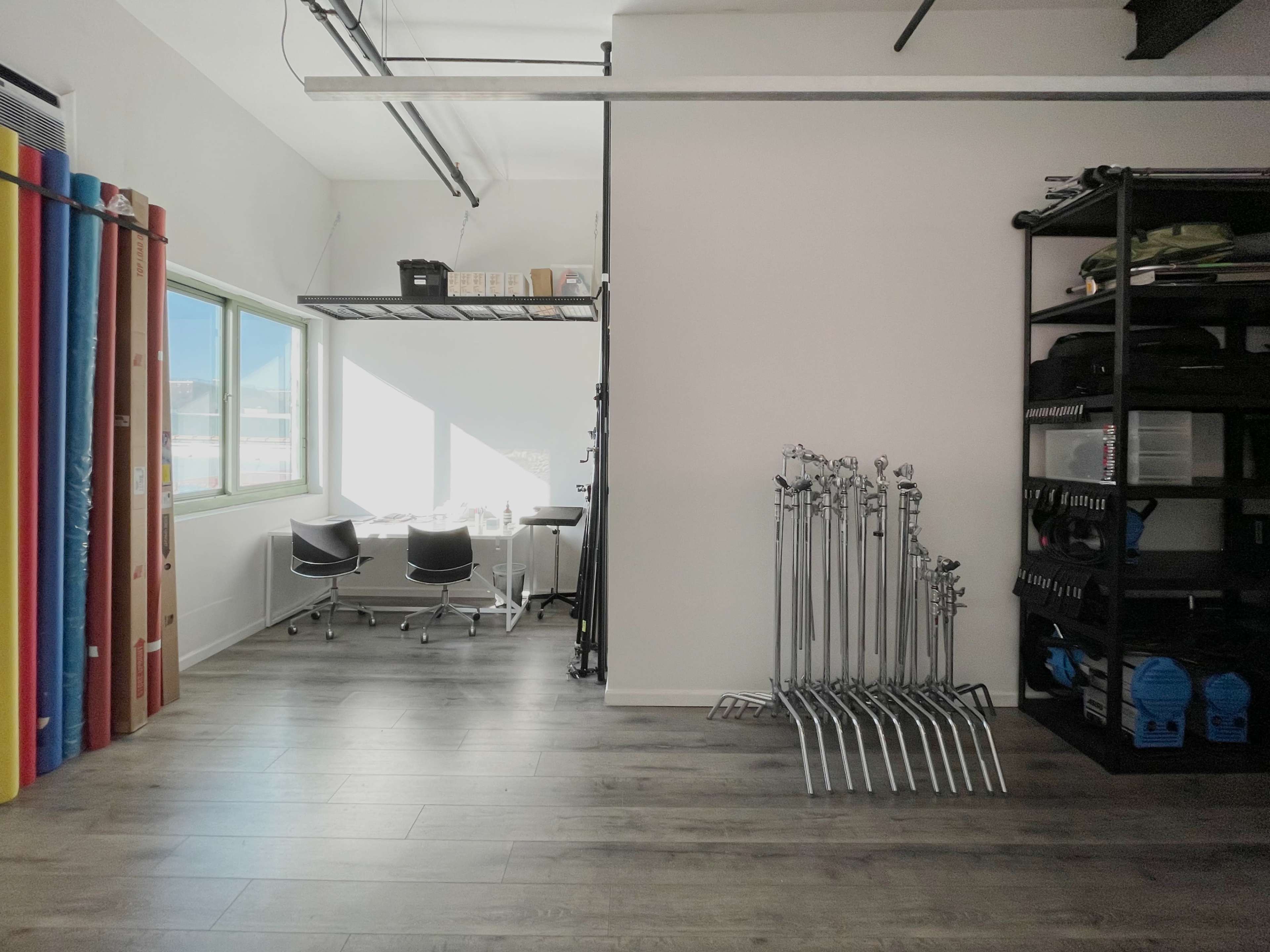 The image shows a spacious room with large windows, featuring a table and chairs on one side and neatly arranged equipment and colorful rolls on the other.