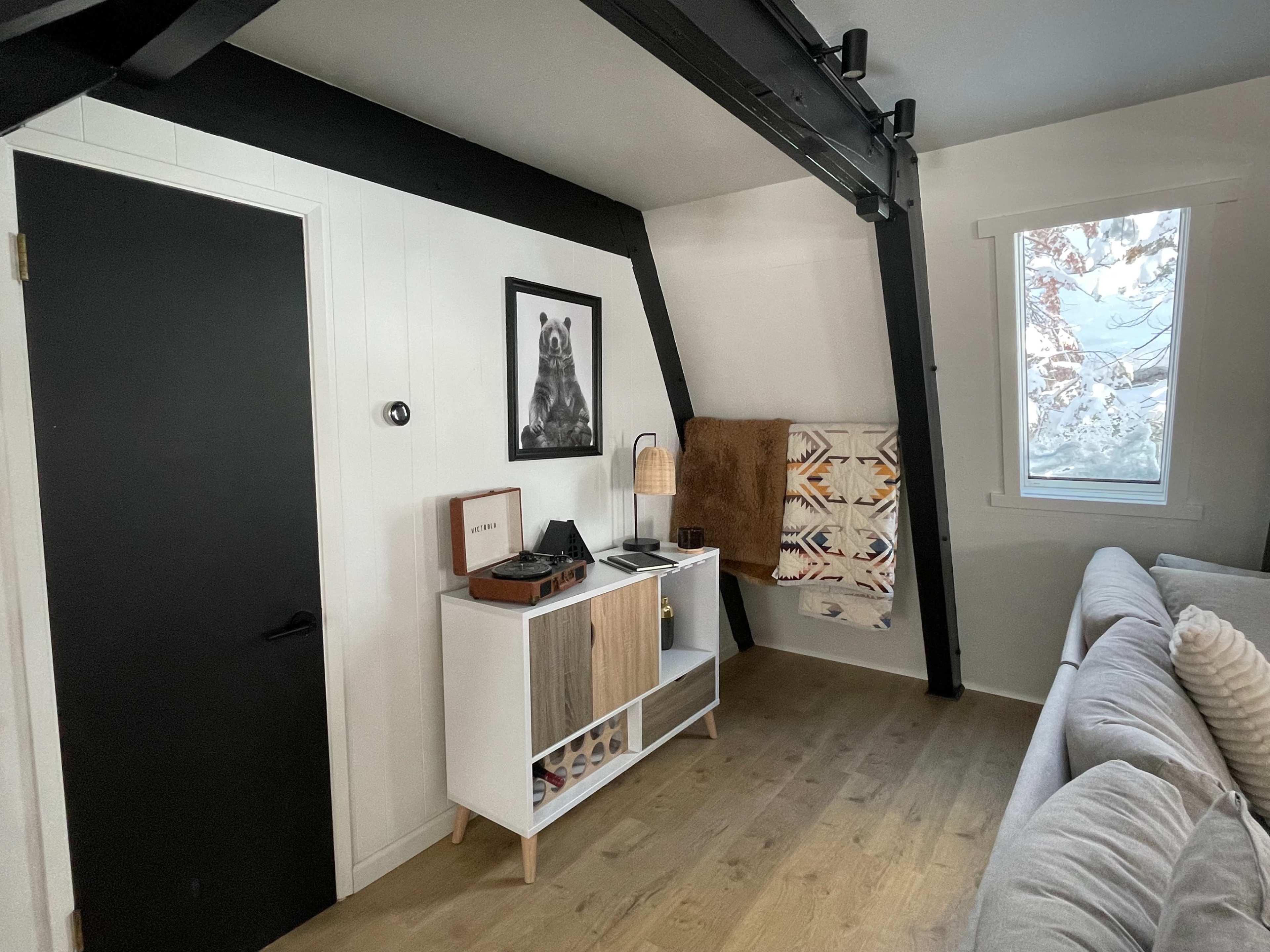 A modern interior with a black door, a white sideboard displaying a record player, and a cozy seating area, complemented by a window and decorative blanket.