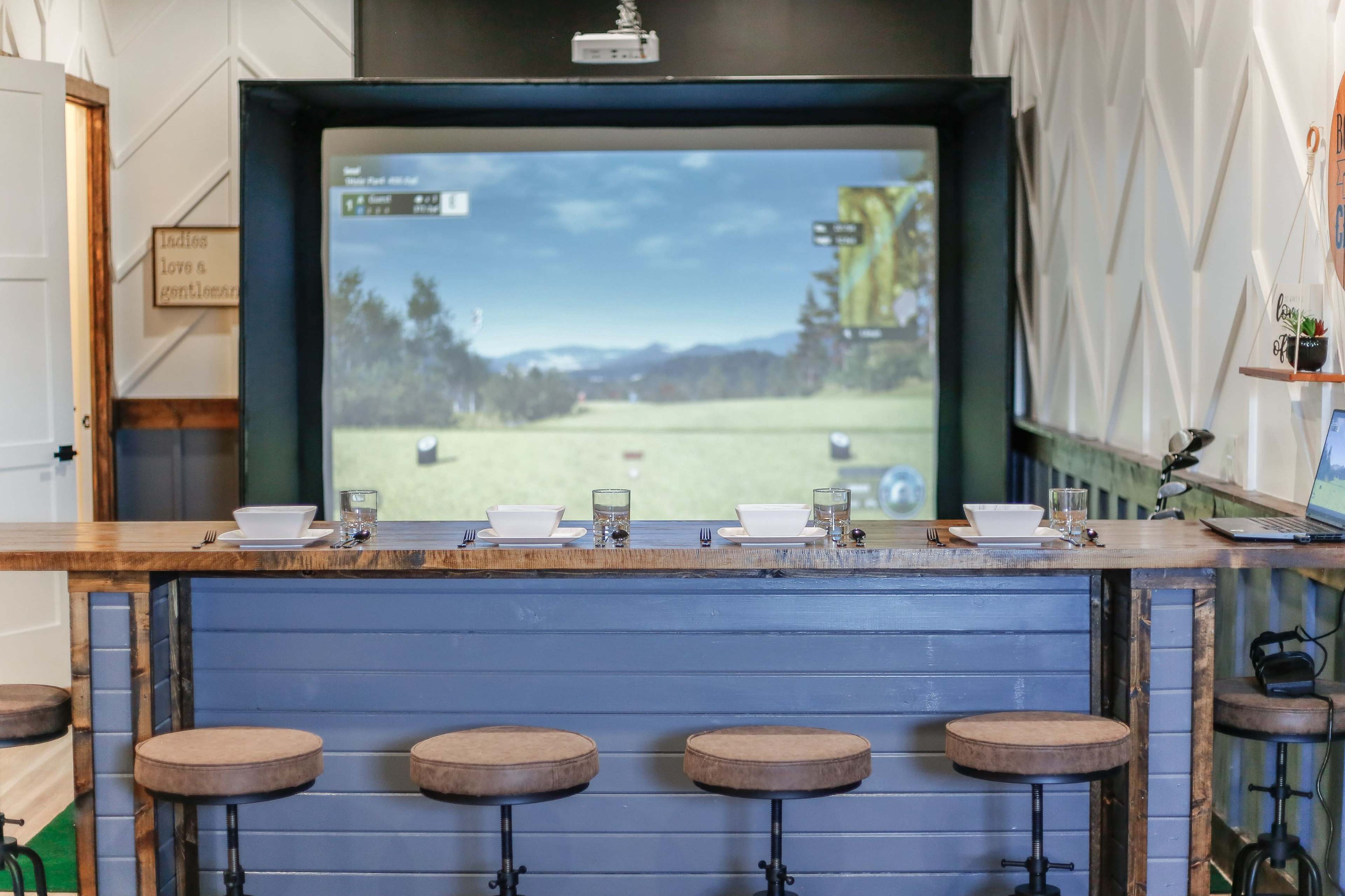 A large projection screen displaying a golf course scene is installed in a room with a bar area featuring stools and glassware.