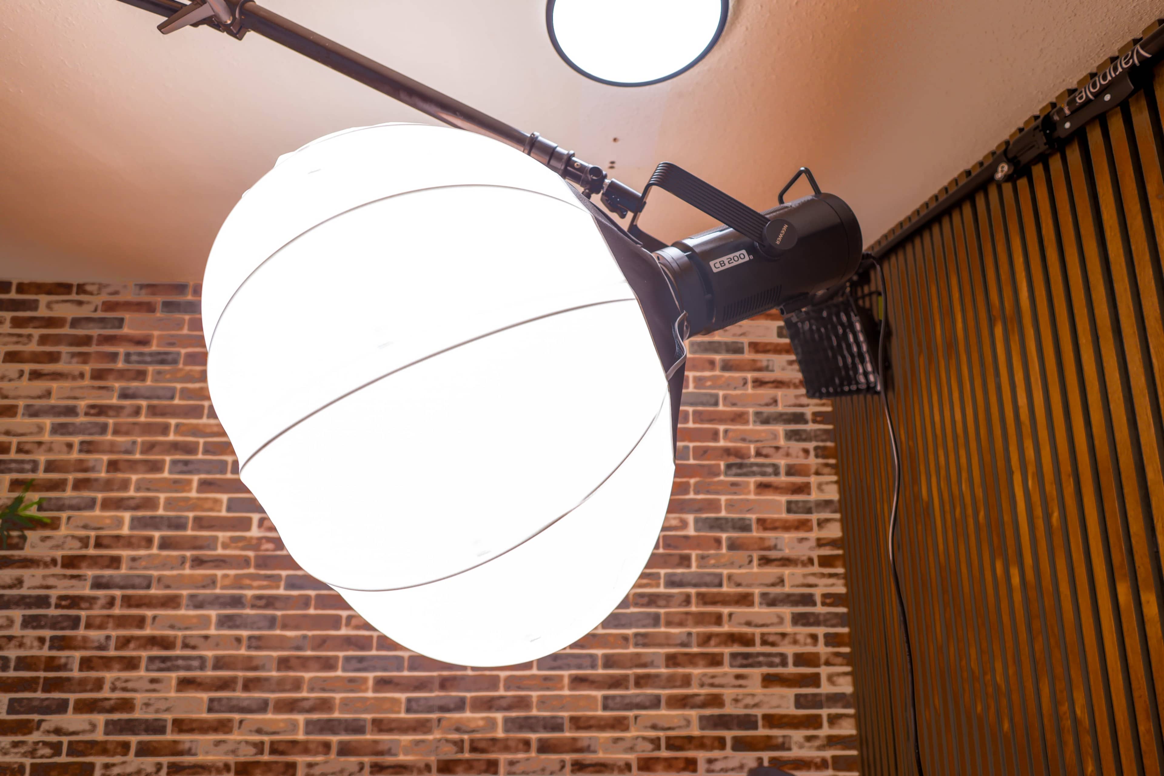 The image shows a large, round light fixture attached to a black arm, mounted on a wall with a brick background.