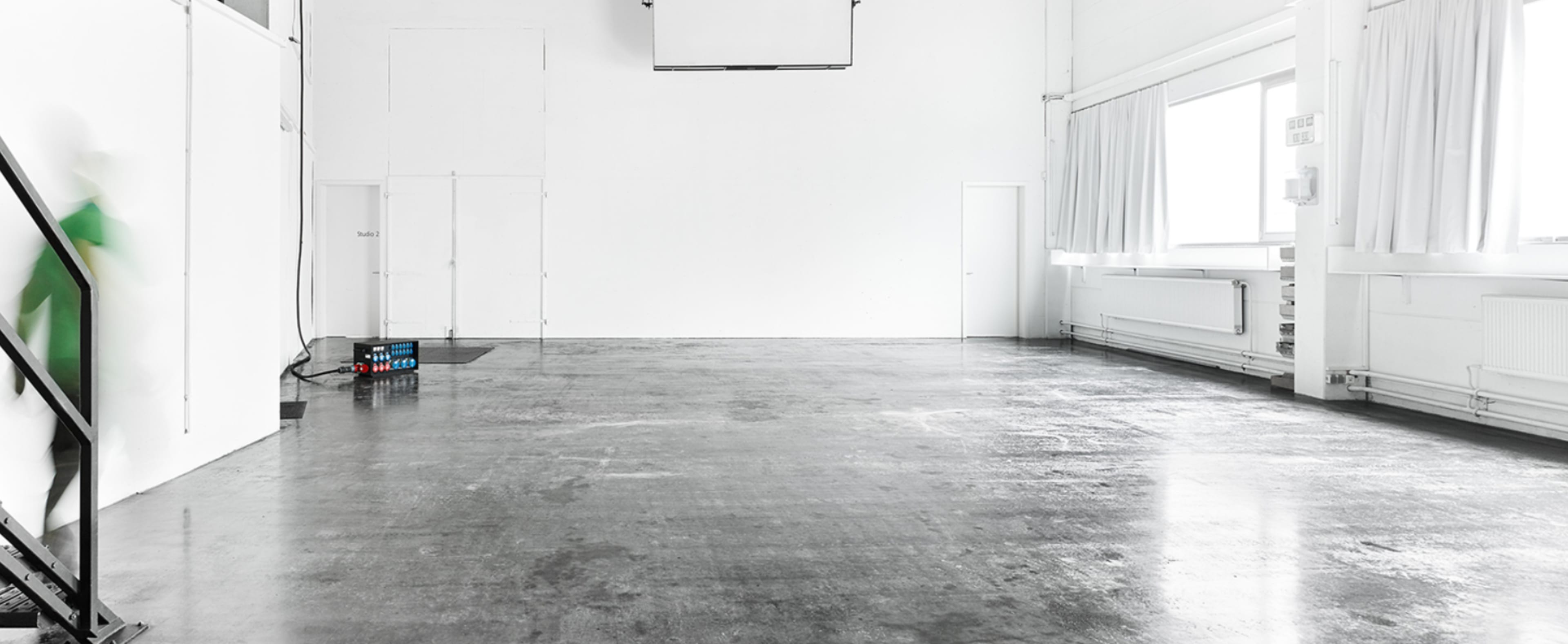 The image shows a spacious, empty room with a polished concrete floor and white walls, featuring a few windows and a staircase leading to a lower level.