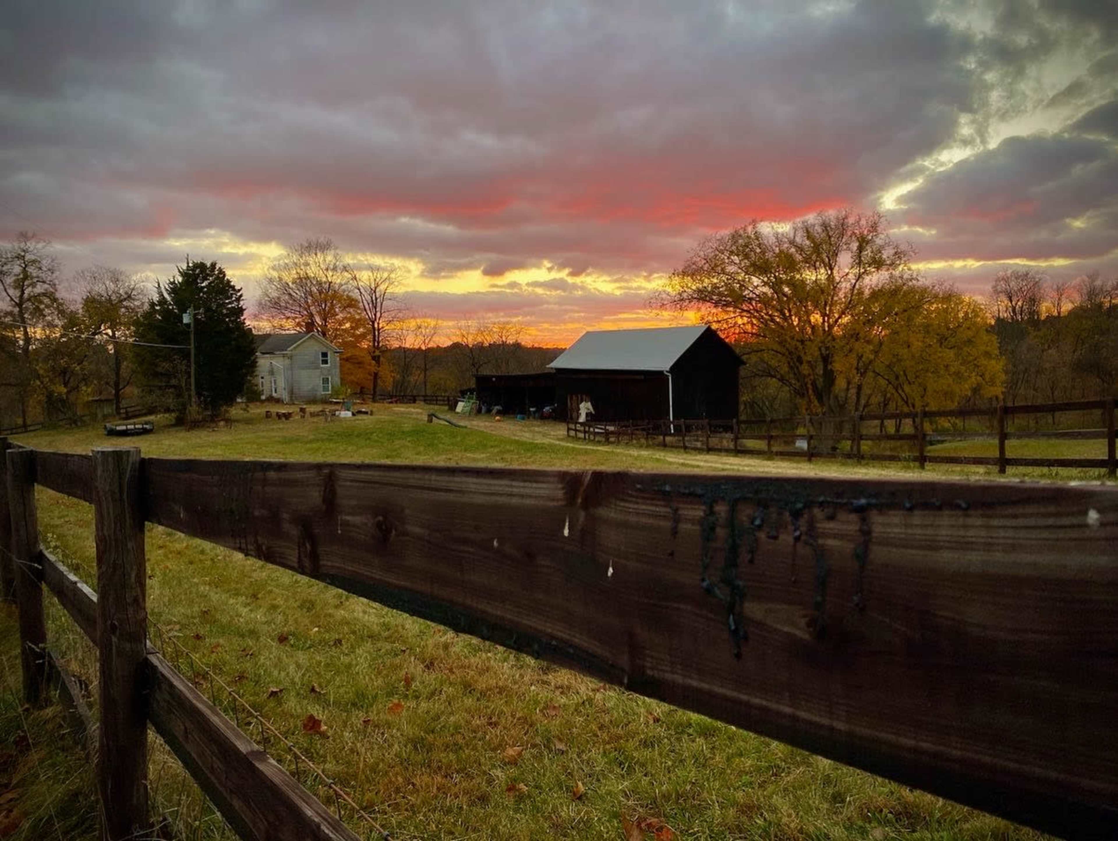 A wooden fence lines a grassy field with a barn and a house silhouetted against a colorful sunset.