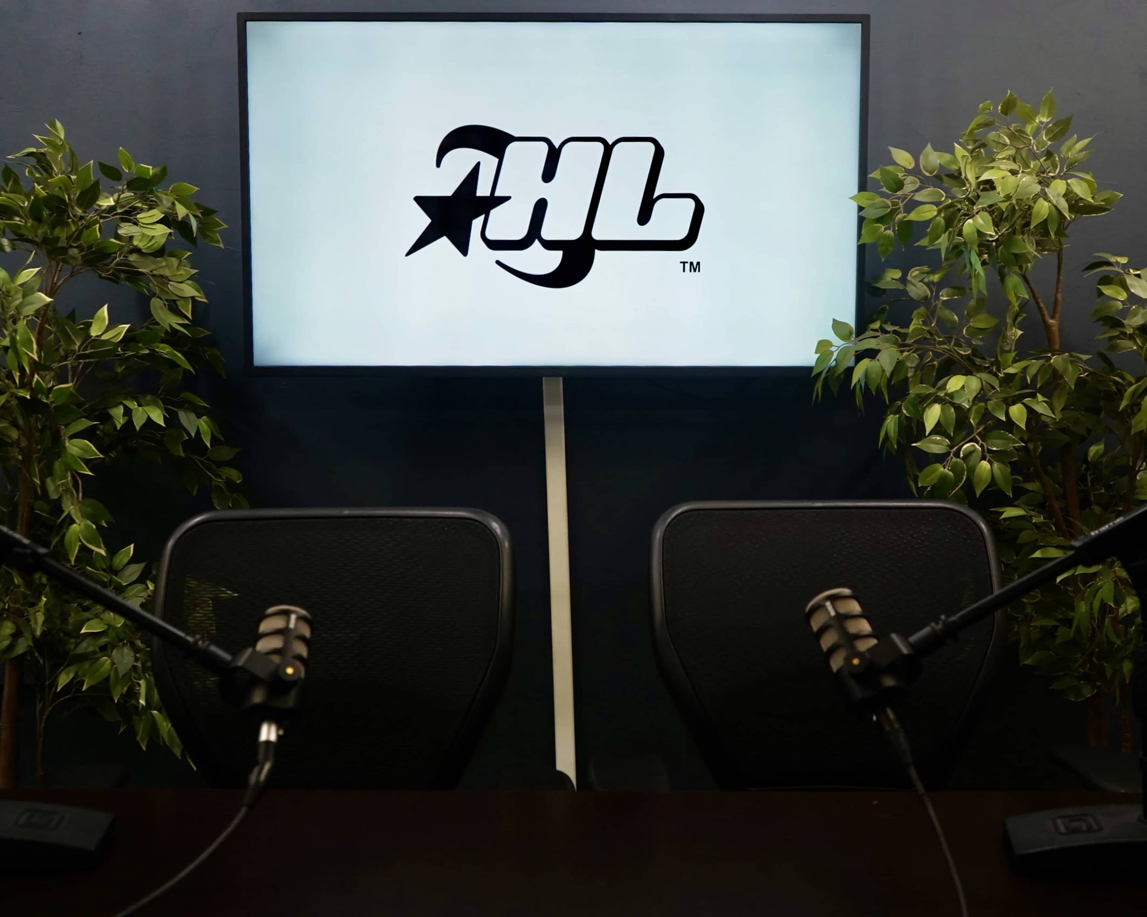 The image shows a recording studio setup with two empty chairs facing a screen displaying a logo, flanked by artificial plants.