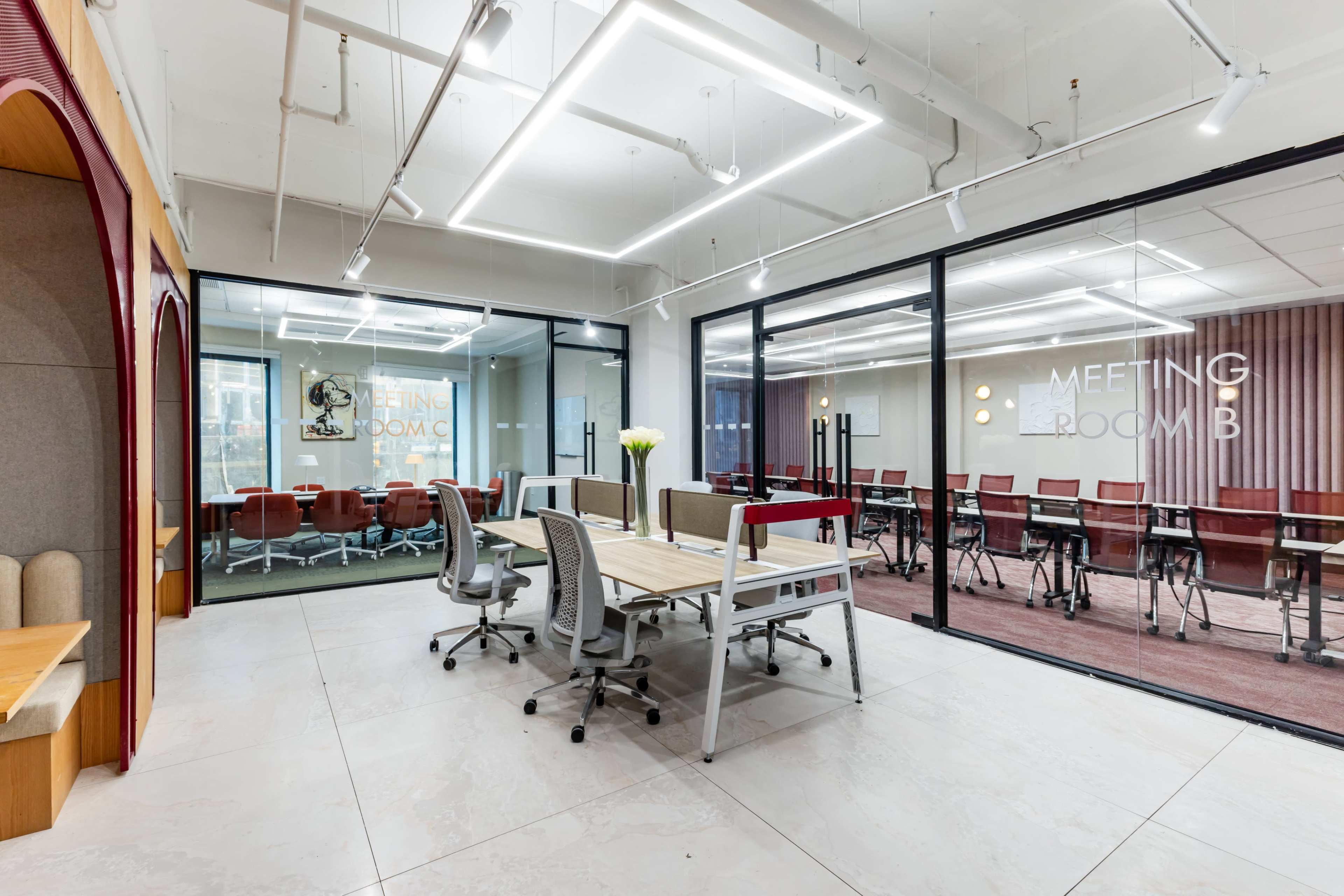 The image shows a modern office space featuring a glass-walled meeting room with seating arrangements and a central table area.