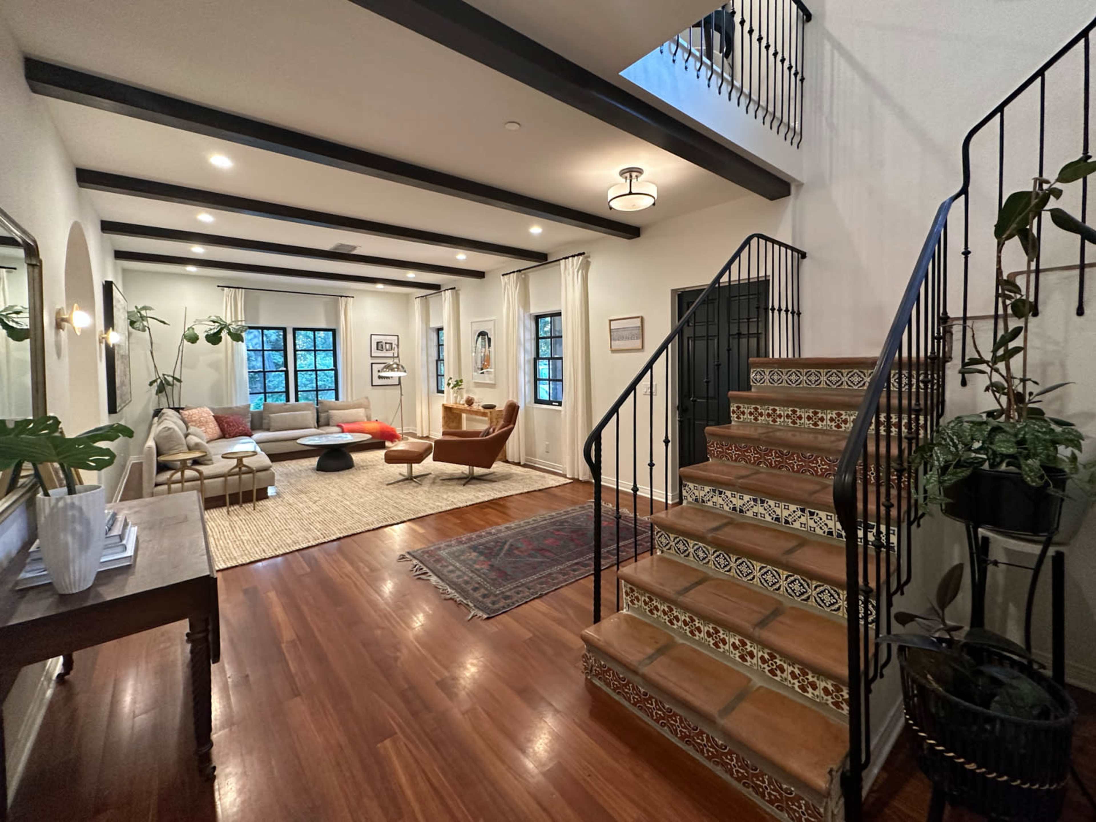 A well-lit living room with a staircase featuring decorative tiles, framed by large windows and furnished with a couch, armchair, and coffee table.