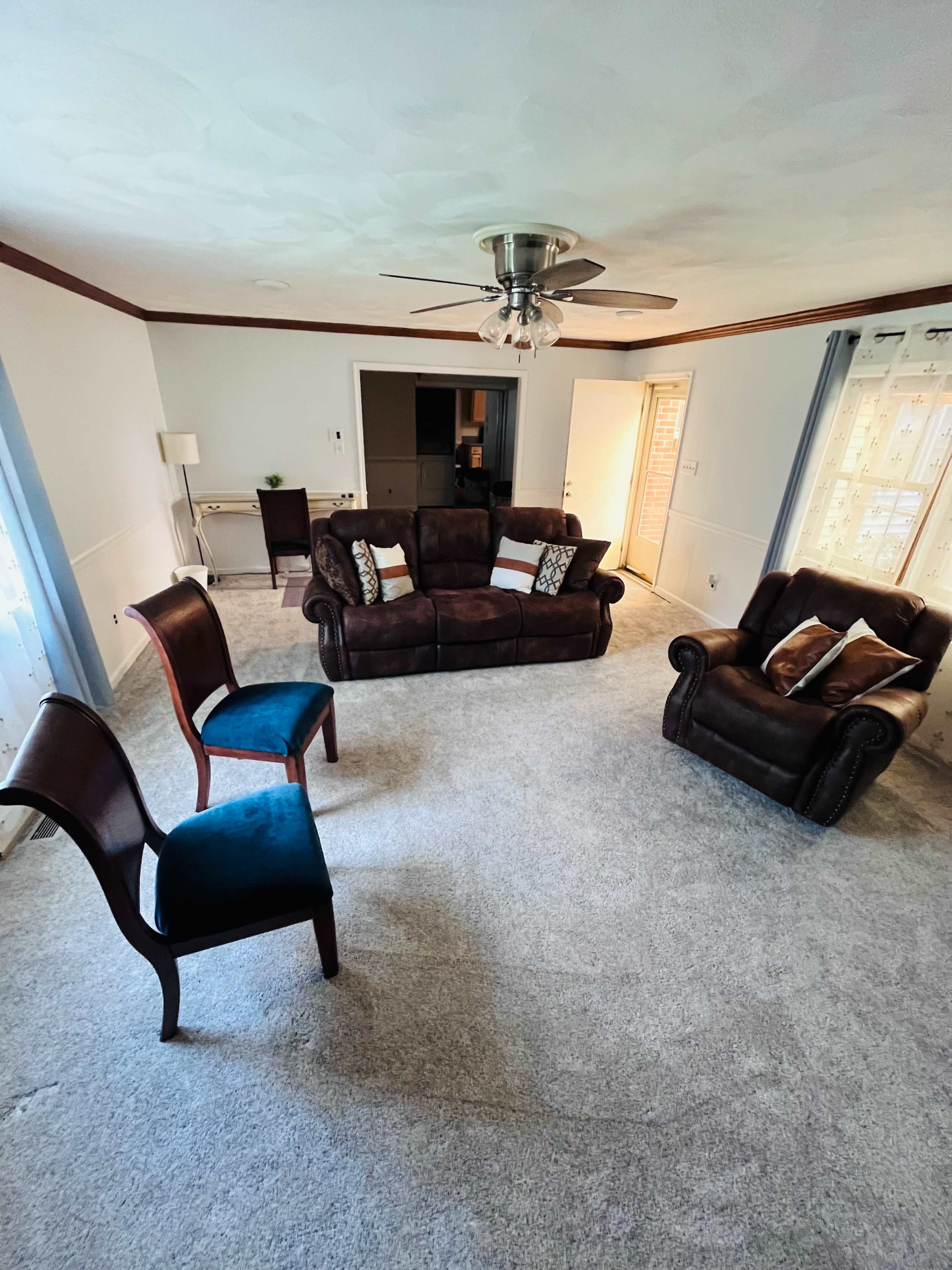 The room features a brown sofa, a brown armchair, and two chairs with one in blue, arranged on a carpeted floor beneath a ceiling fan.