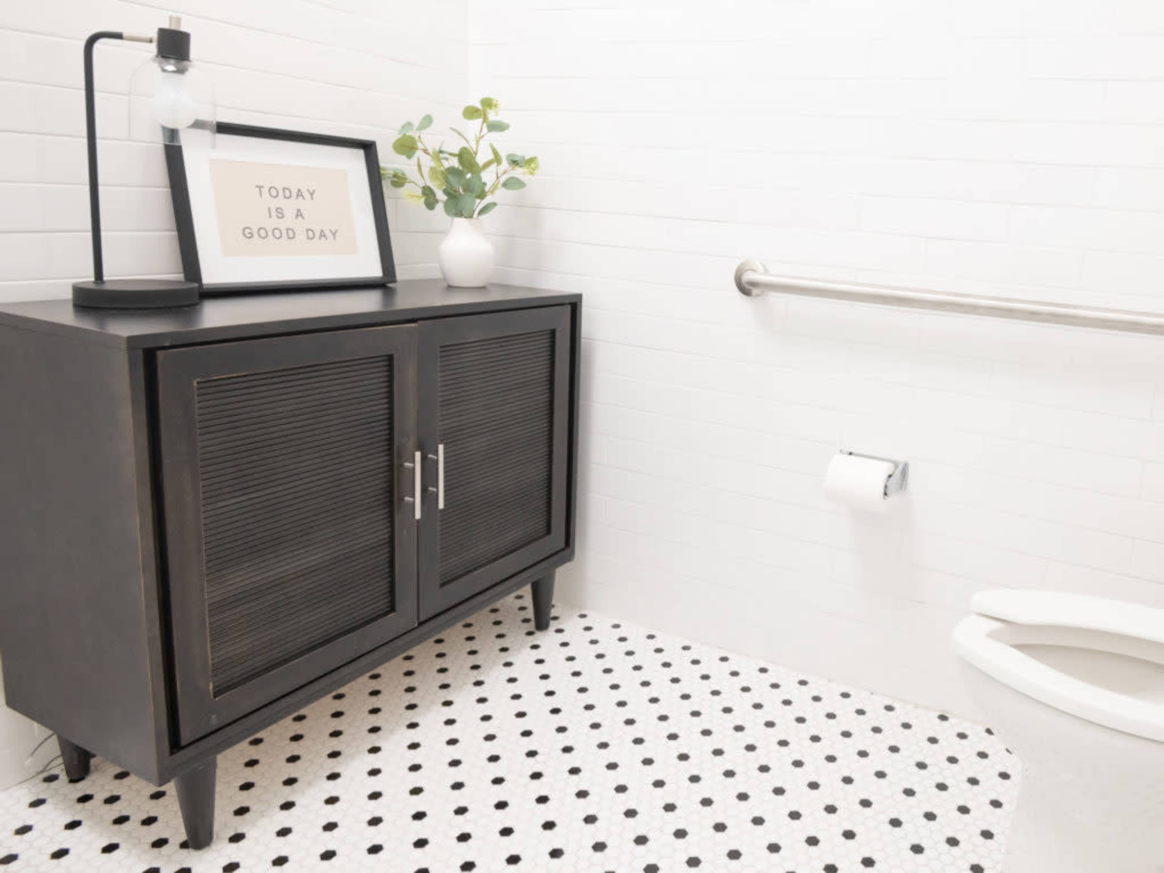A bathroom corner with a black cabinet, a framed sign on top, a small plant, and a toilet on a polka-dot floor.