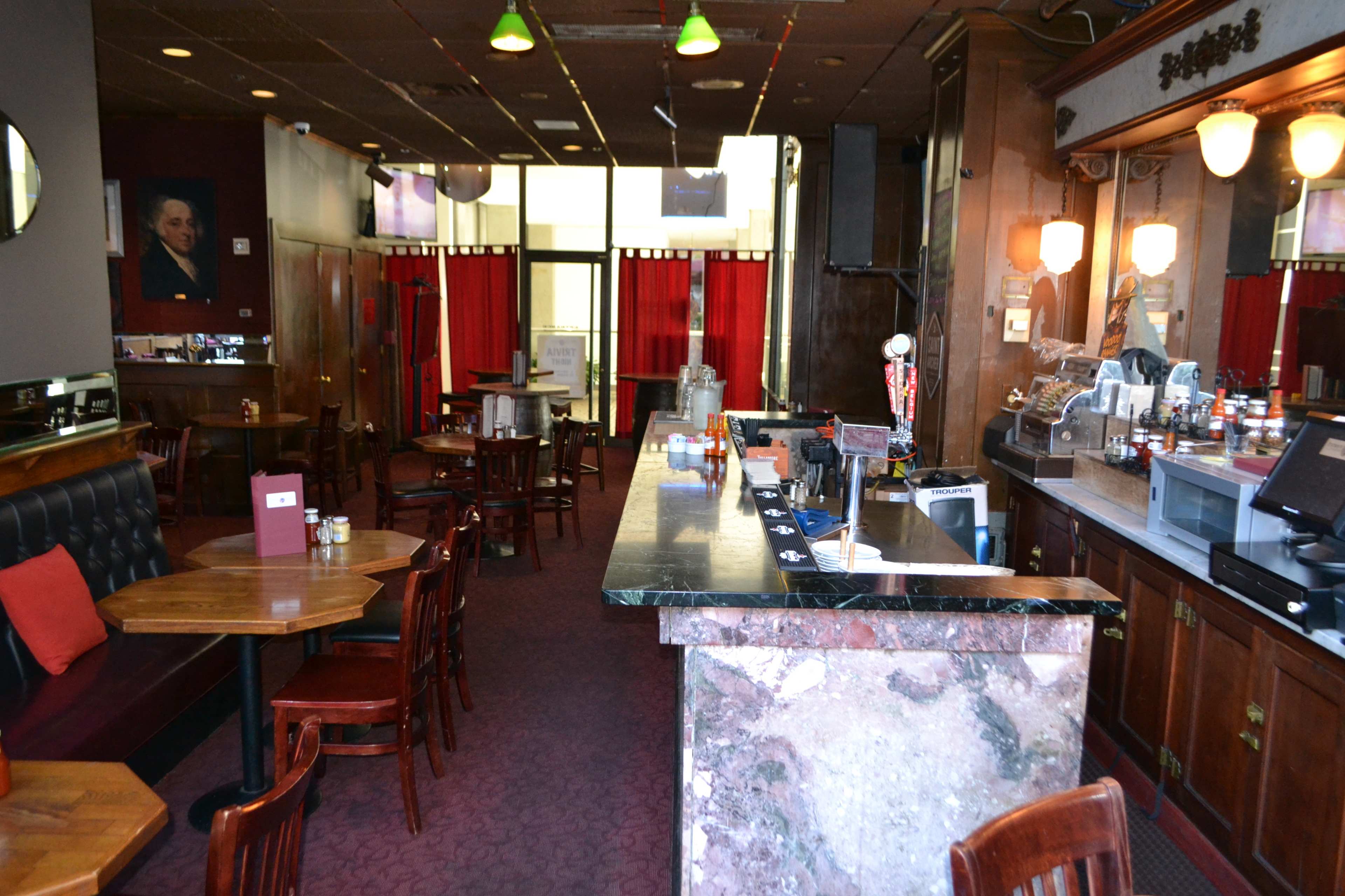 An empty restaurant interior featuring wooden tables and chairs, a bar with a marble counter, and red curtains in the background.
