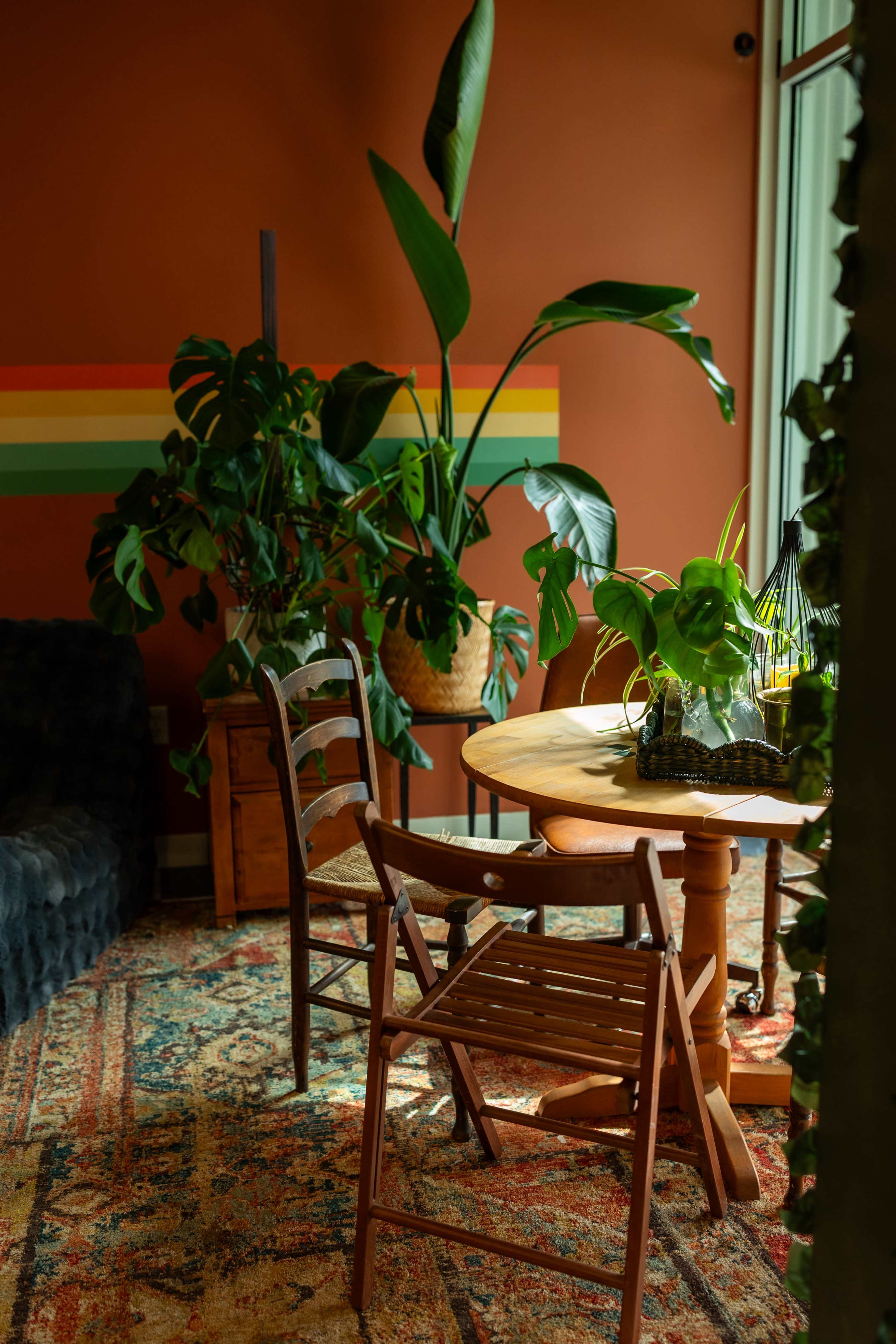 A dining area features a round wooden table surrounded by chairs, with plants and colorful wall stripes adding to the decor.