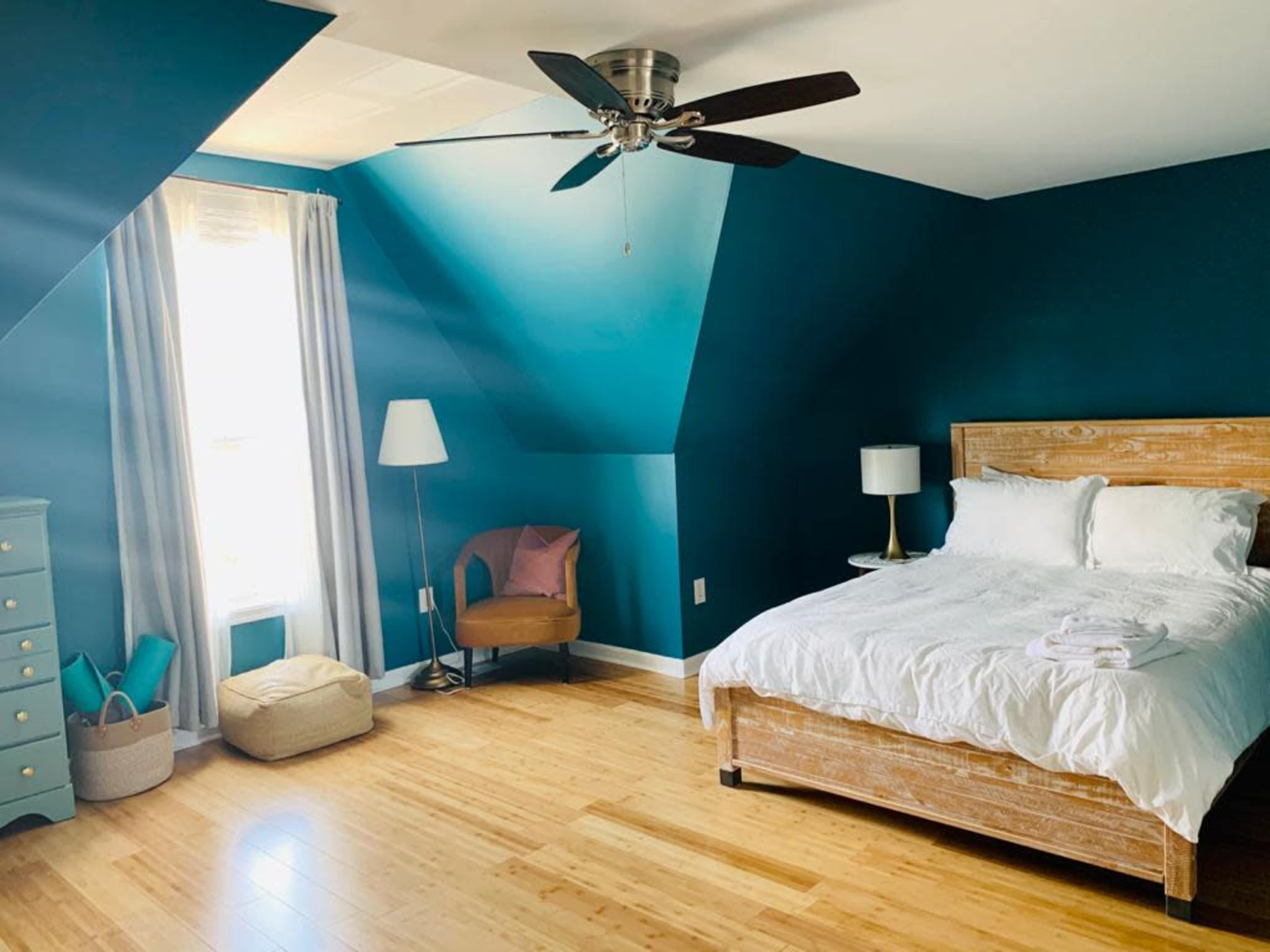 A bright bedroom features teal walls, a wooden bed with white linens, a chair, a lamp, and bamboo flooring.
