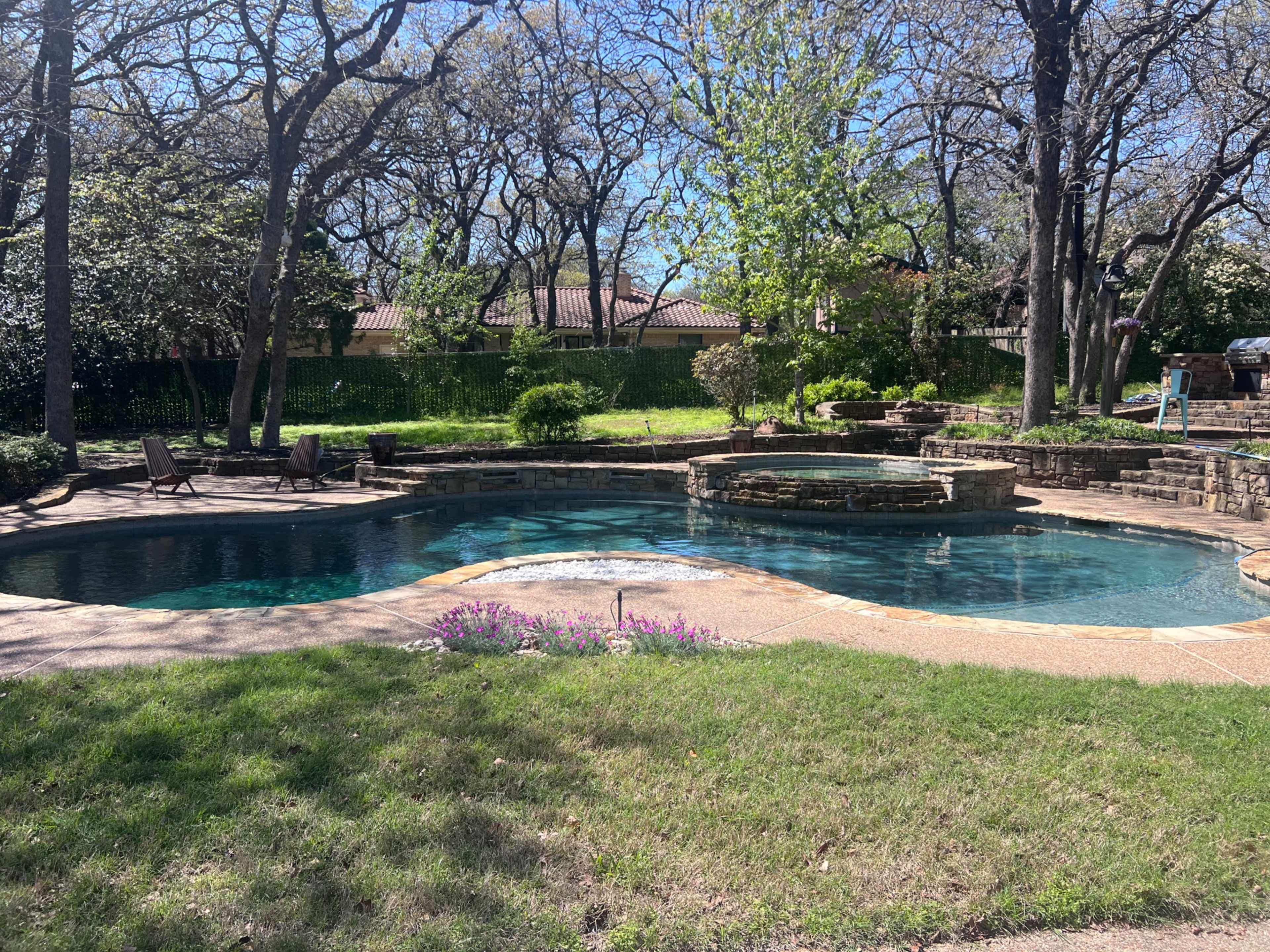 The image shows an outdoor swimming pool with a stone hot tub surrounded by trees and grass.