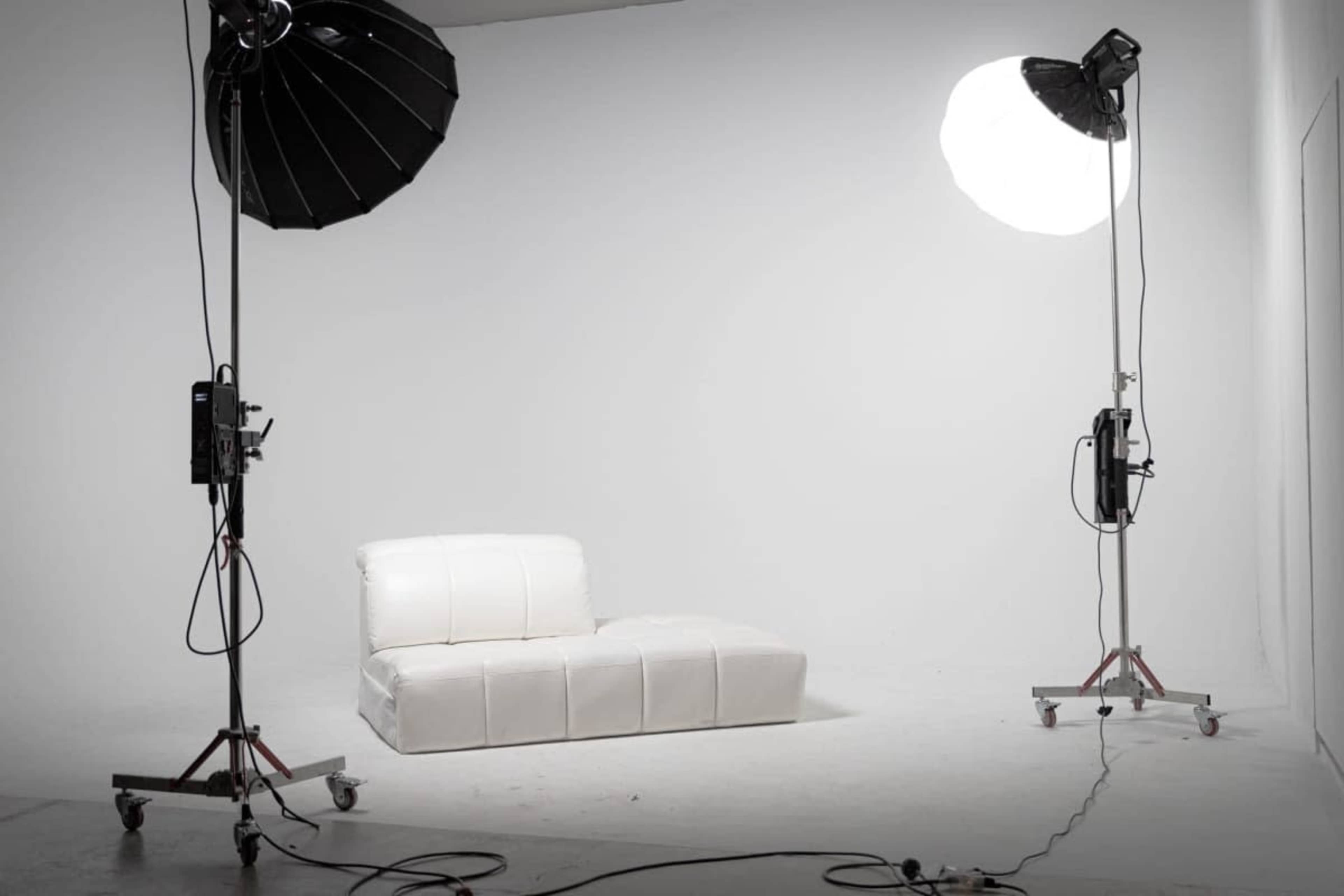 The image shows a minimalist photography studio with a white backdrop, featuring a single white couch illuminated by two large studio lights.