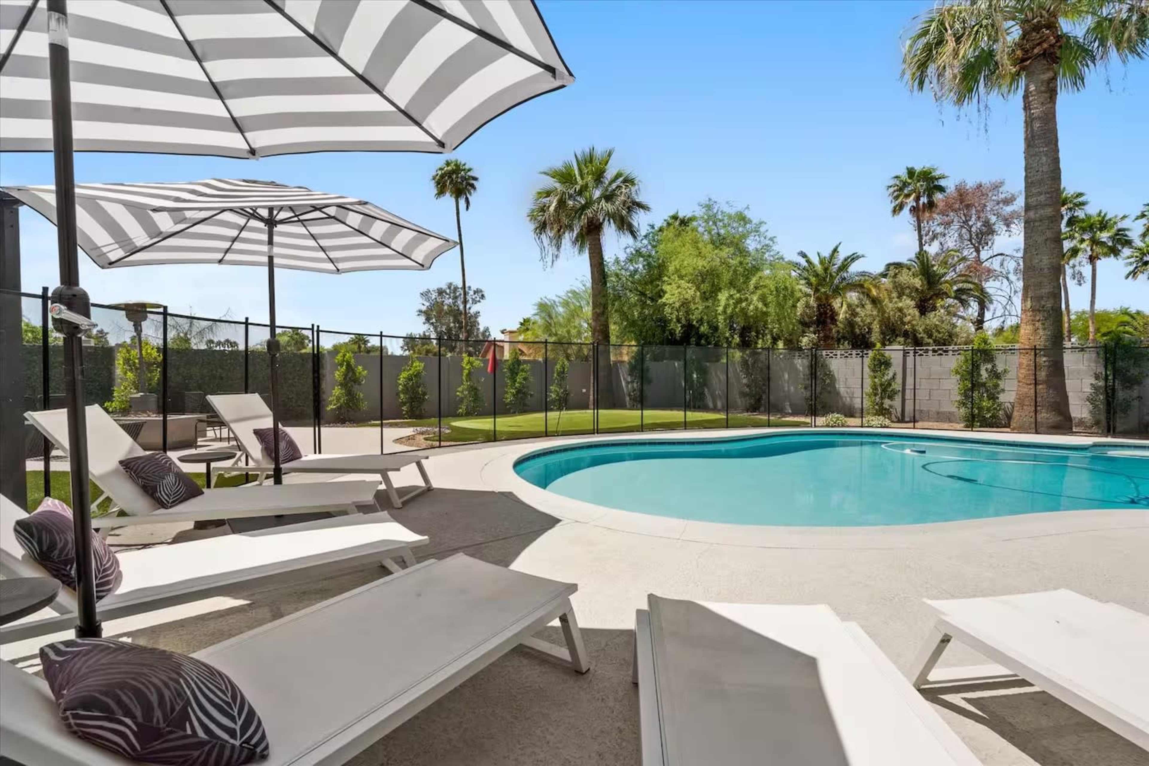 A swimming pool surrounded by lounge chairs and umbrellas, with palm trees and greenery in the background.