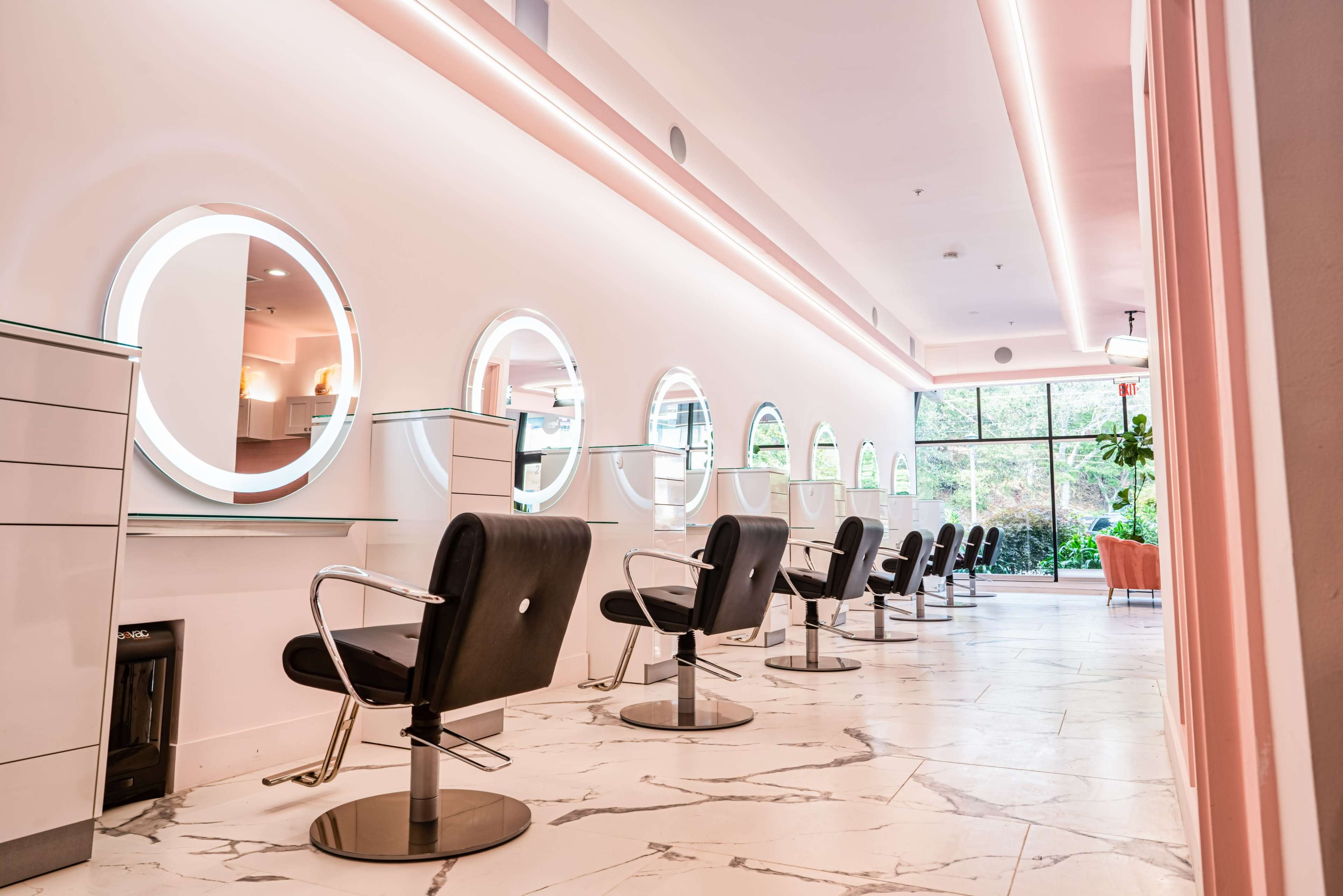 A modern hair salon with multiple styling stations featuring round mirrors and black salon chairs, lined along a bright, airy space.