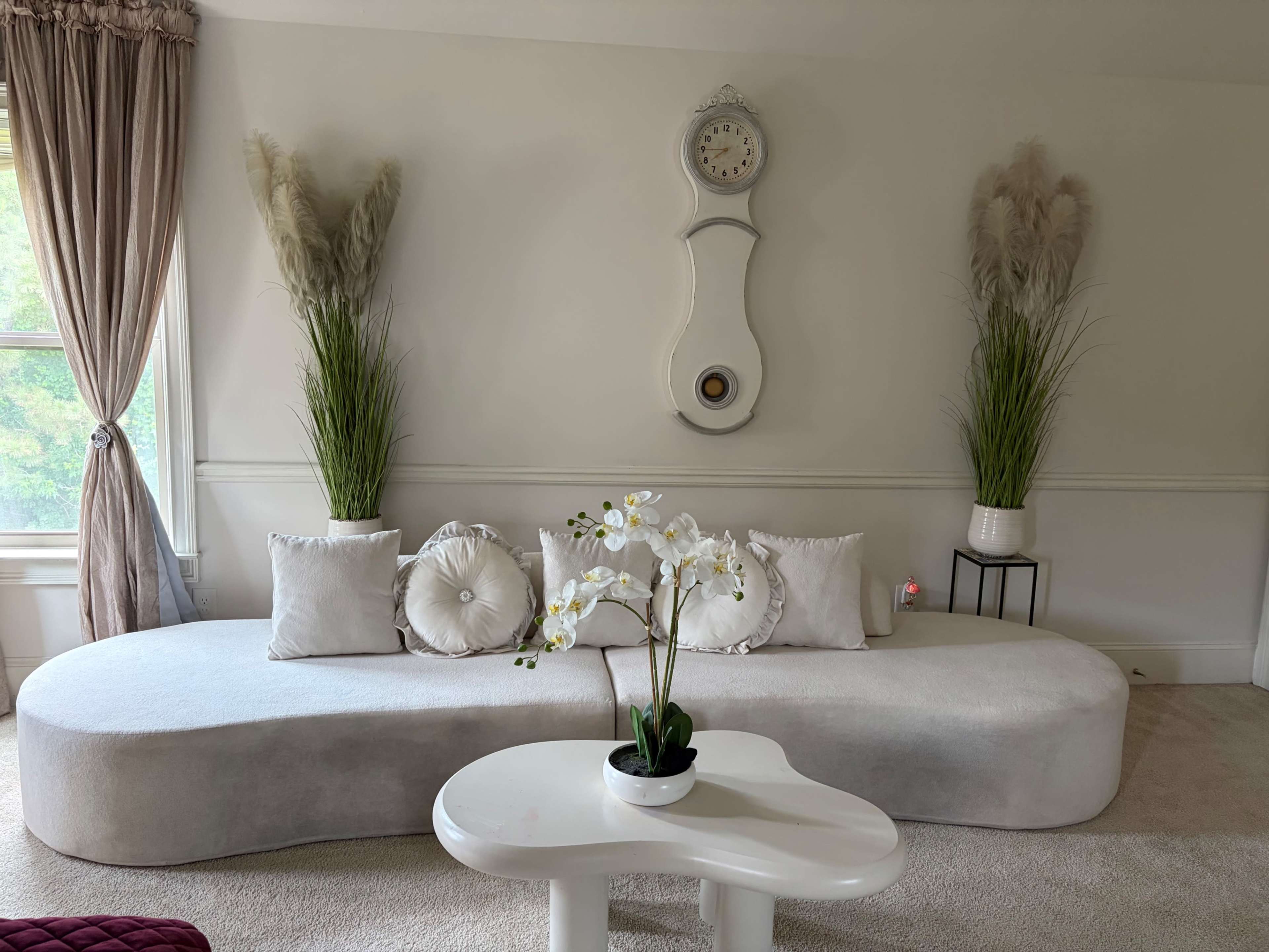 The image shows a modern living room featuring a curved gray sofa with decorative pillows and a white table, flanked by tall plants and a wall clock above.