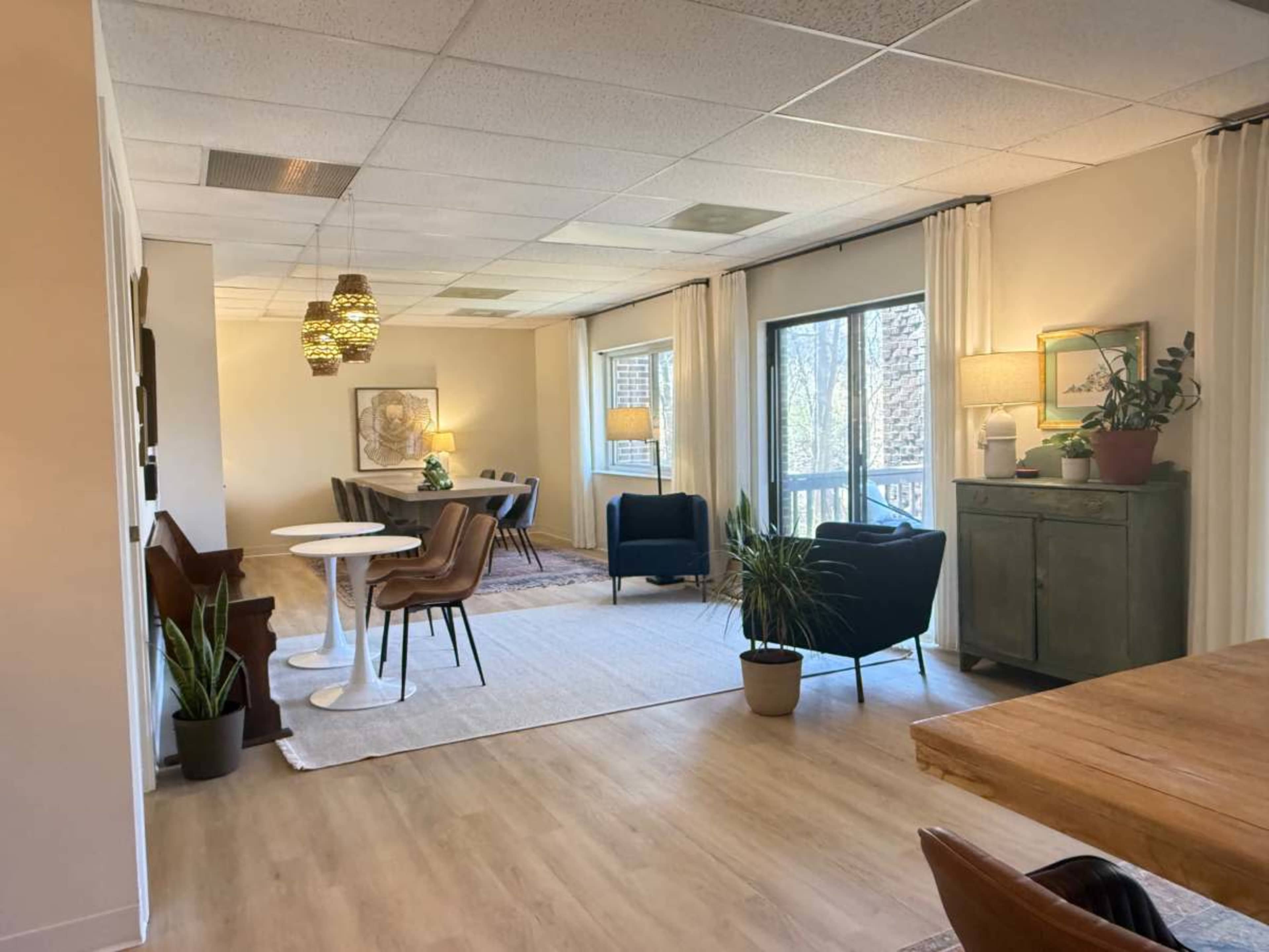 The image depicts a spacious, well-lit room with a mix of seating areas, including a dining table, chairs, and a green cabinet, surrounded by large windows.