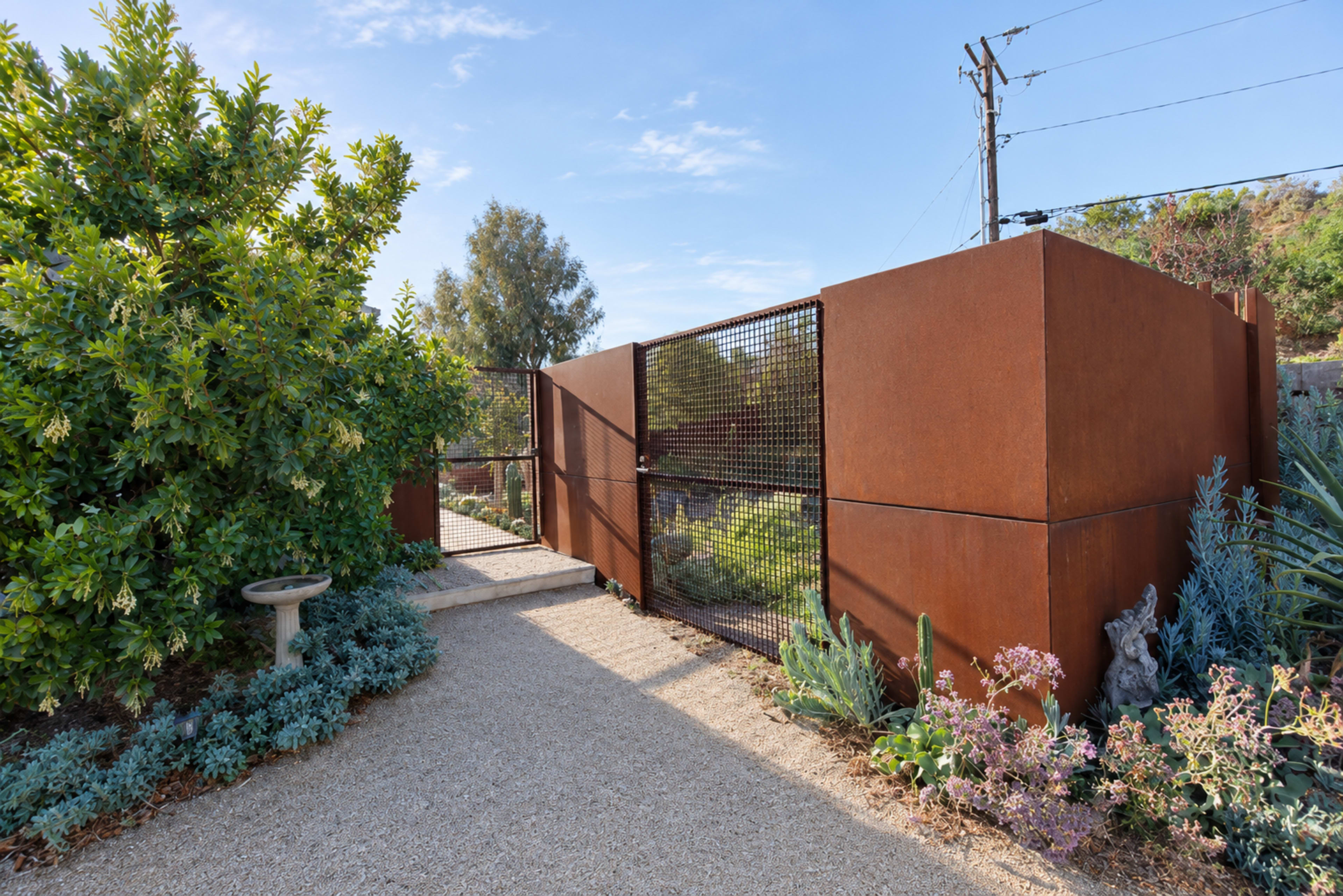 The image shows a pathway leading to a metal gate surrounded by various plants and greenery.
