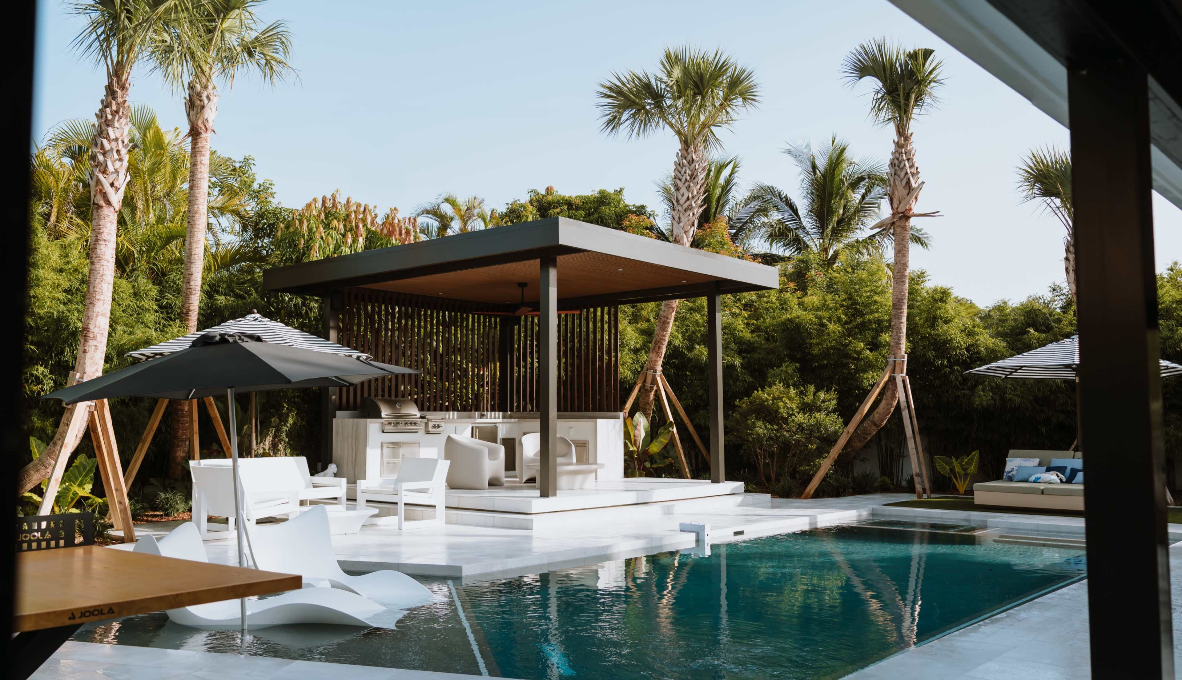 A modern outdoor pool area features a sleek pavilion, lounge chairs, and palm trees under a clear blue sky.