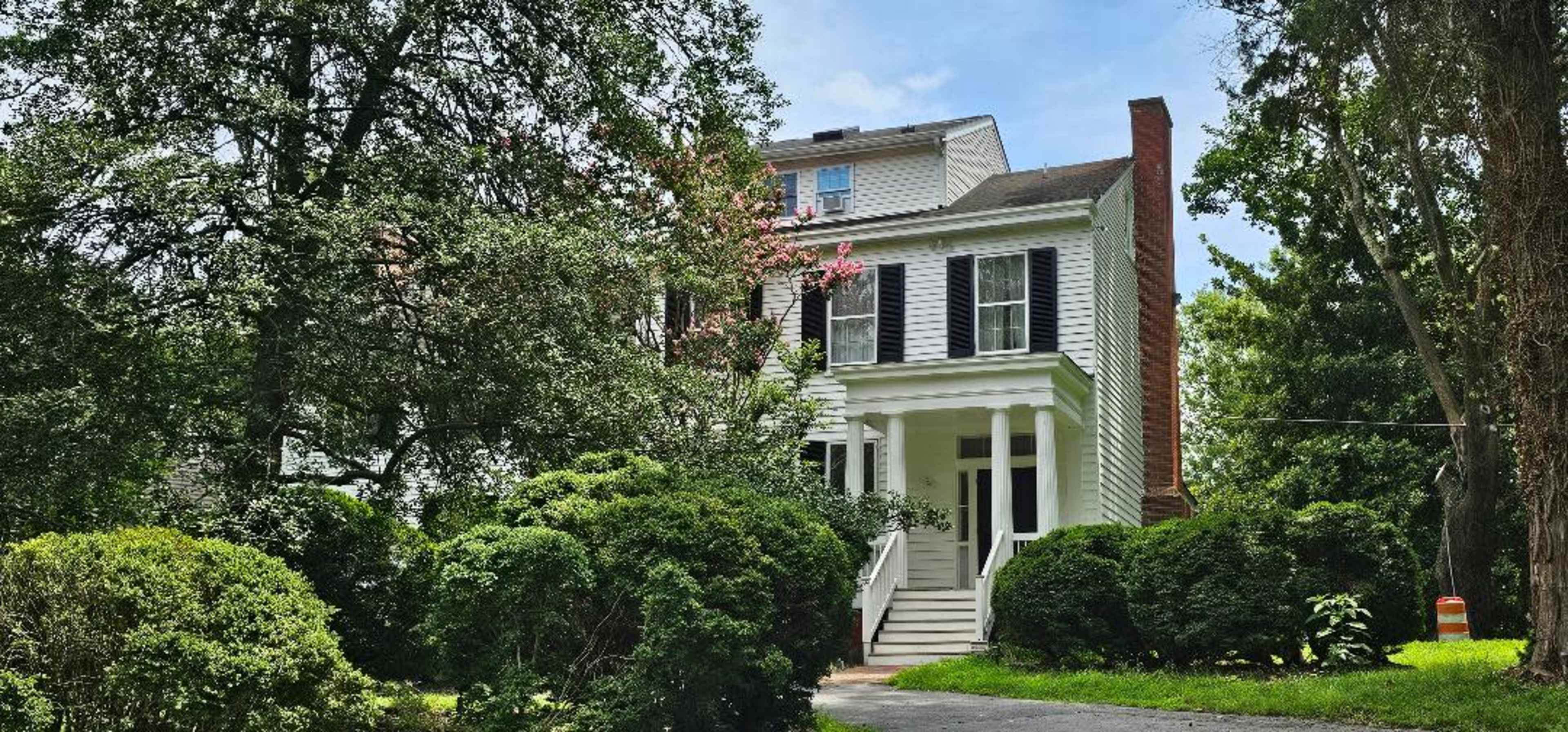 A multi-story white house with black shutters, surrounded by lush green foliage and trees.