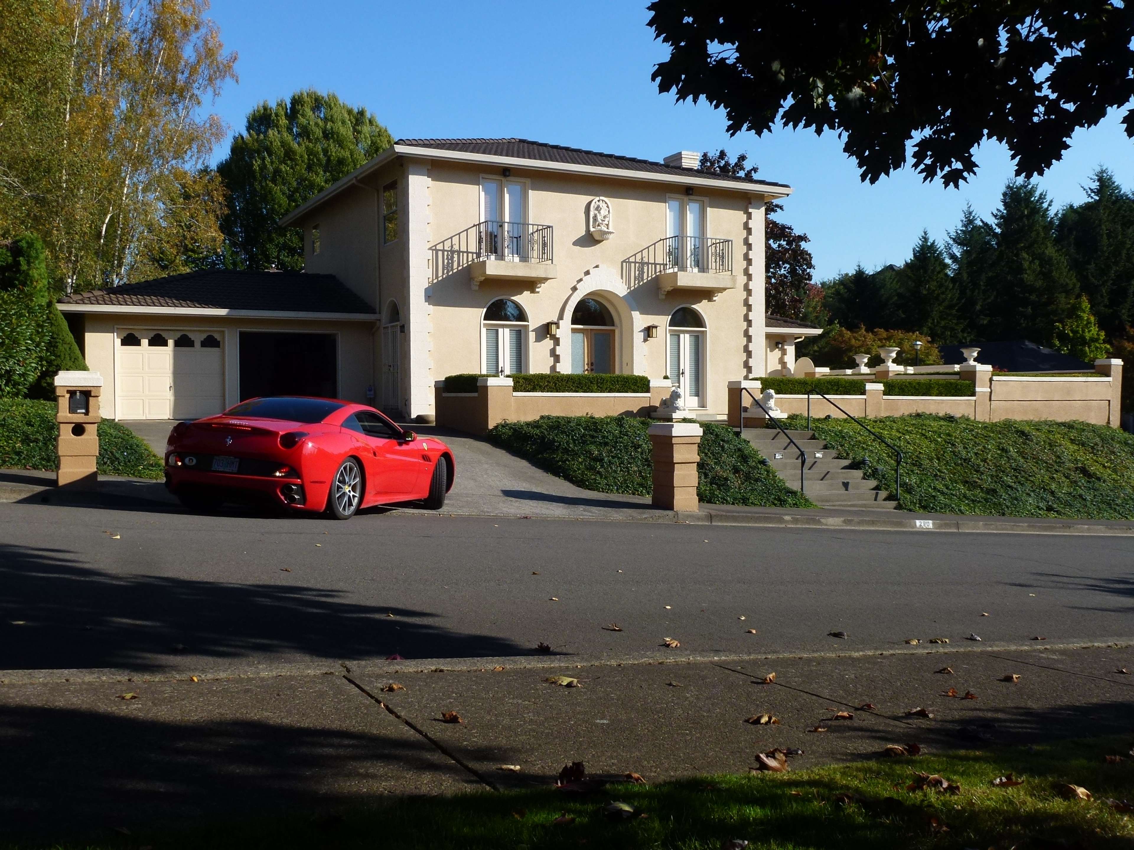 A red sports car is parked in front of a large, two-story house with a landscaped yard and a curved driveway.