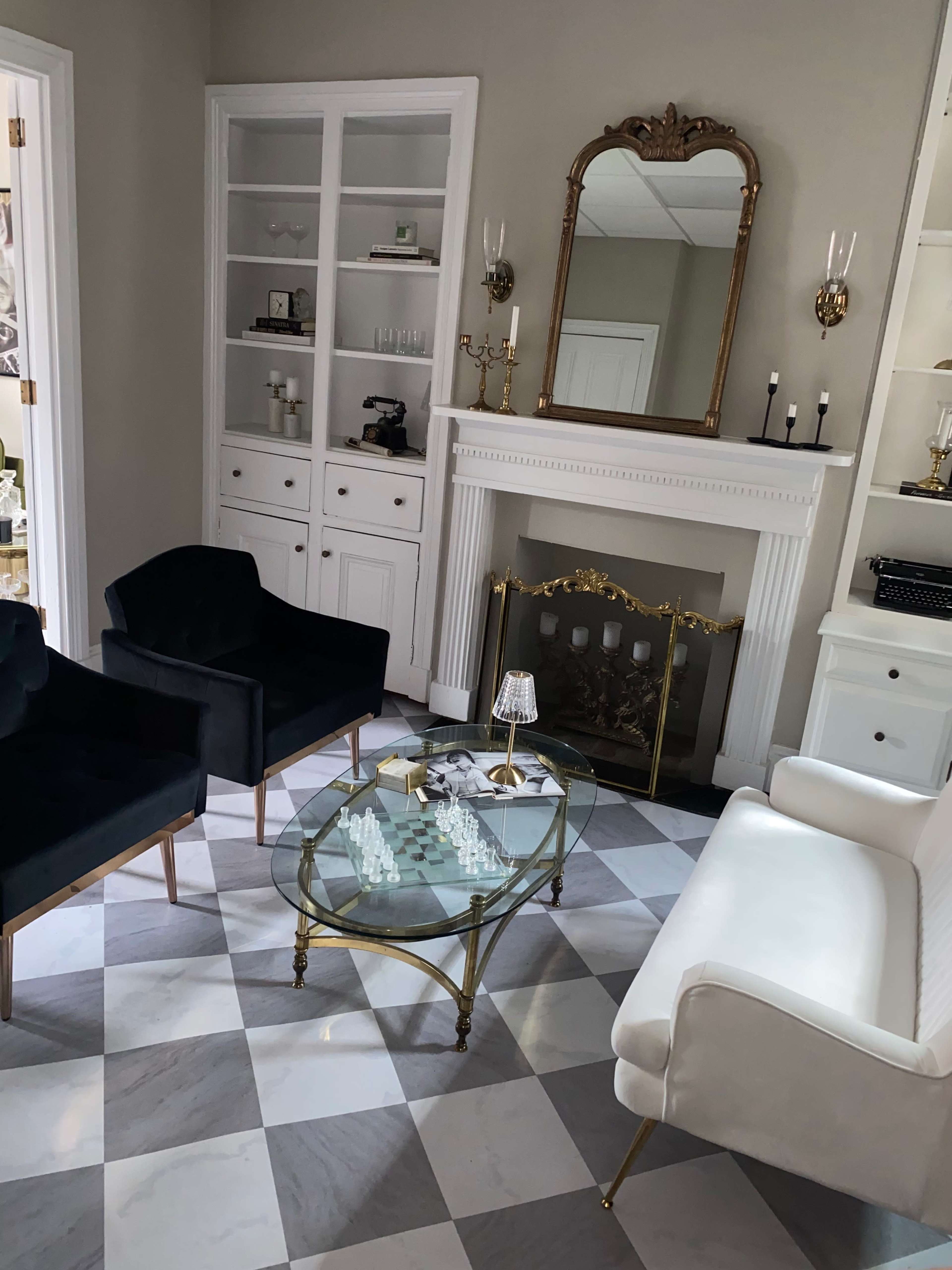 A living room features a glass coffee table with a chess set, flanked by two black armchairs and a white couch, with a fireplace and built-in shelves in the background.