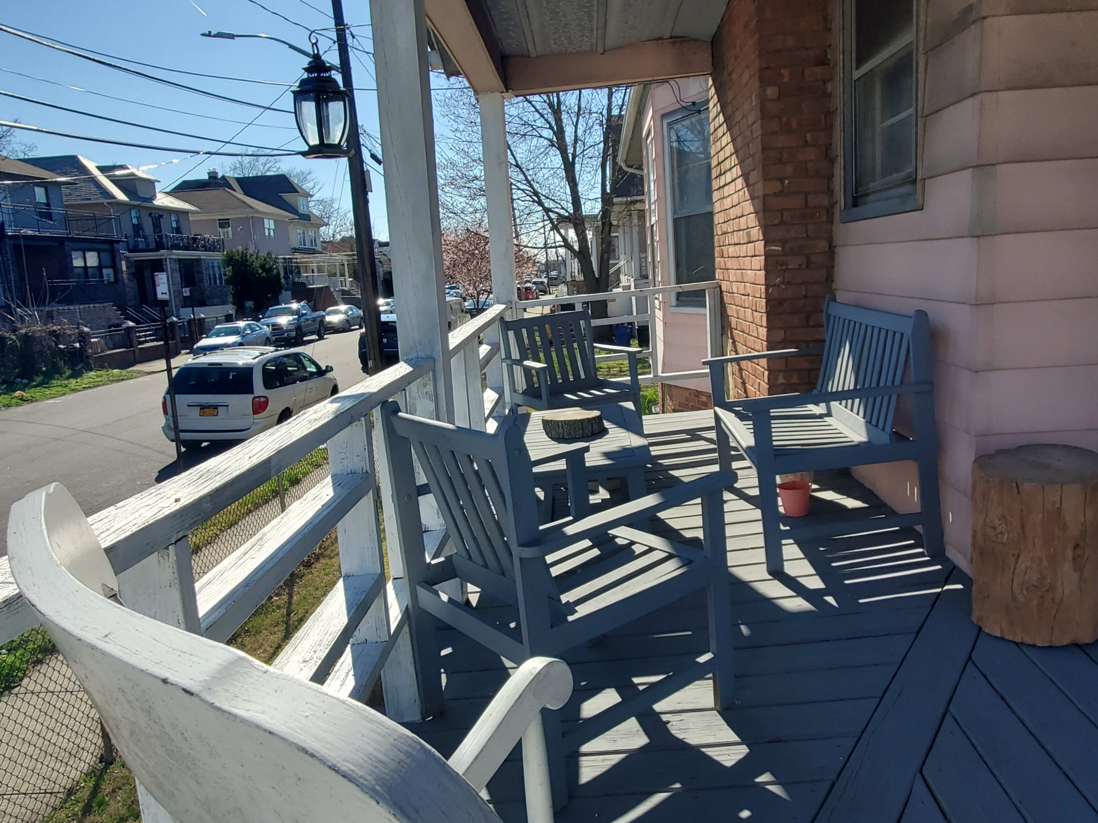 A porch with gray wooden chairs and a table, overlooking a street lined with parked cars and houses.