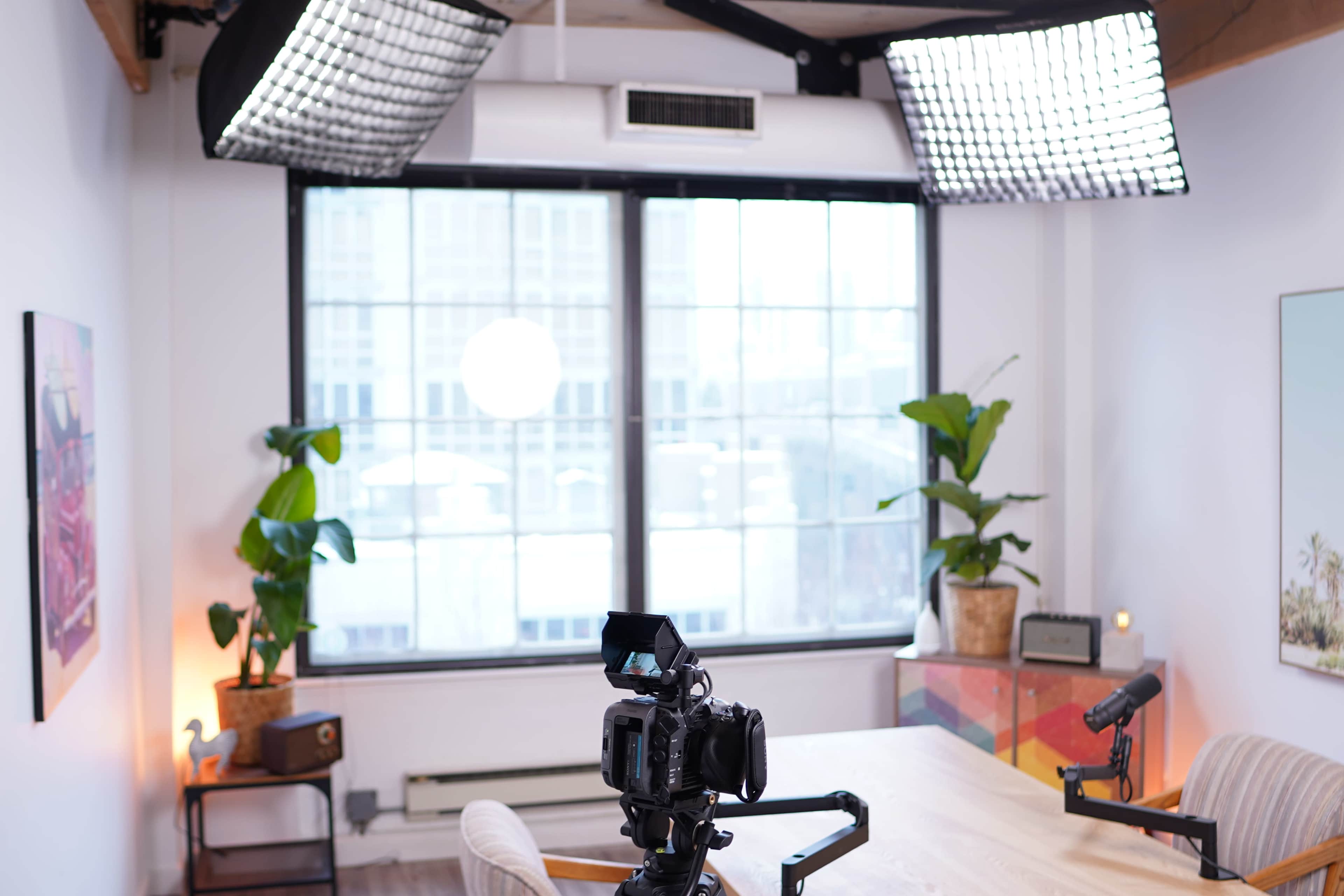 A well-lit room features a camera on a tripod facing a table, with large windows displaying greenery outside.
